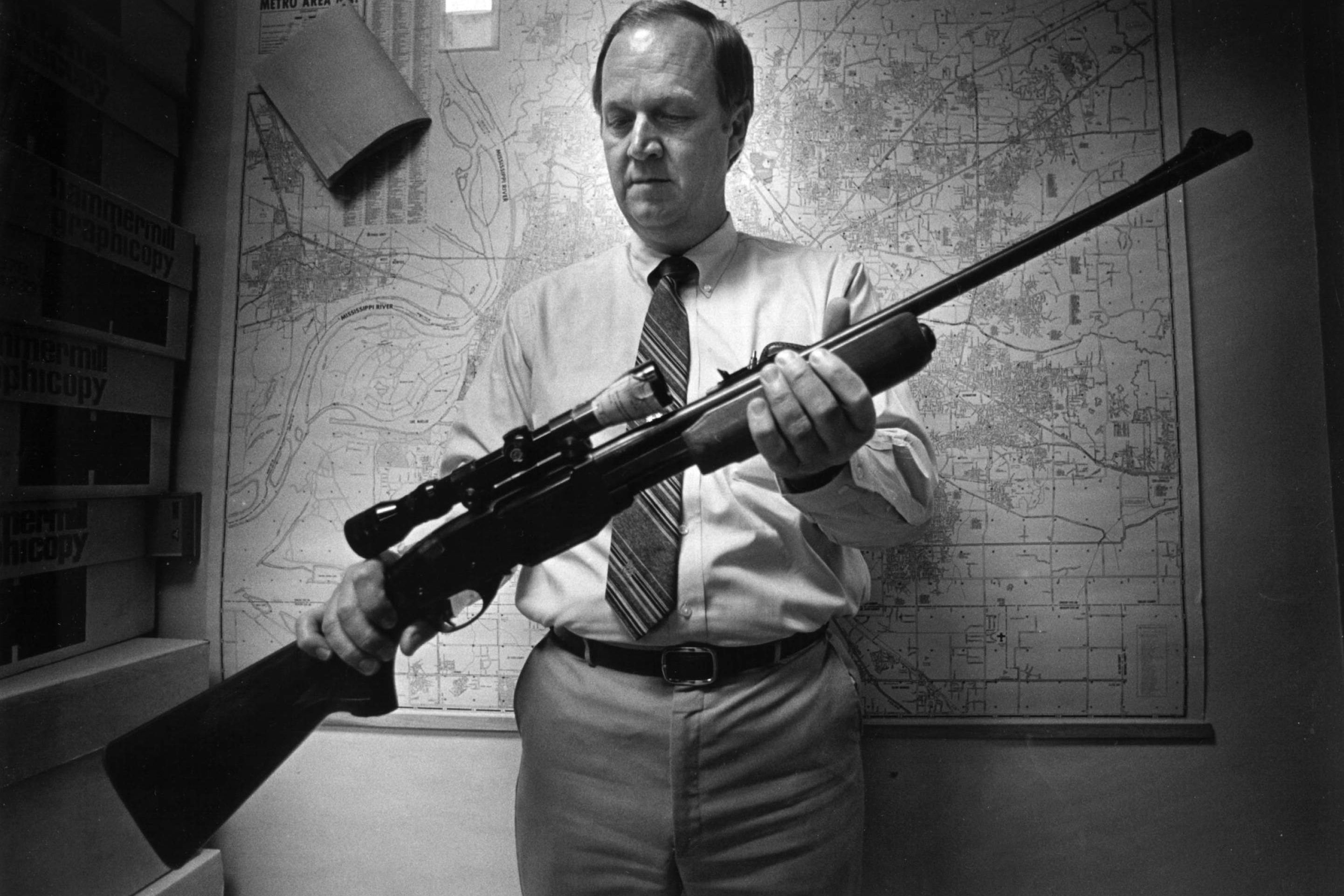 A middle-aged white man displaying a rifle in front of a large map.