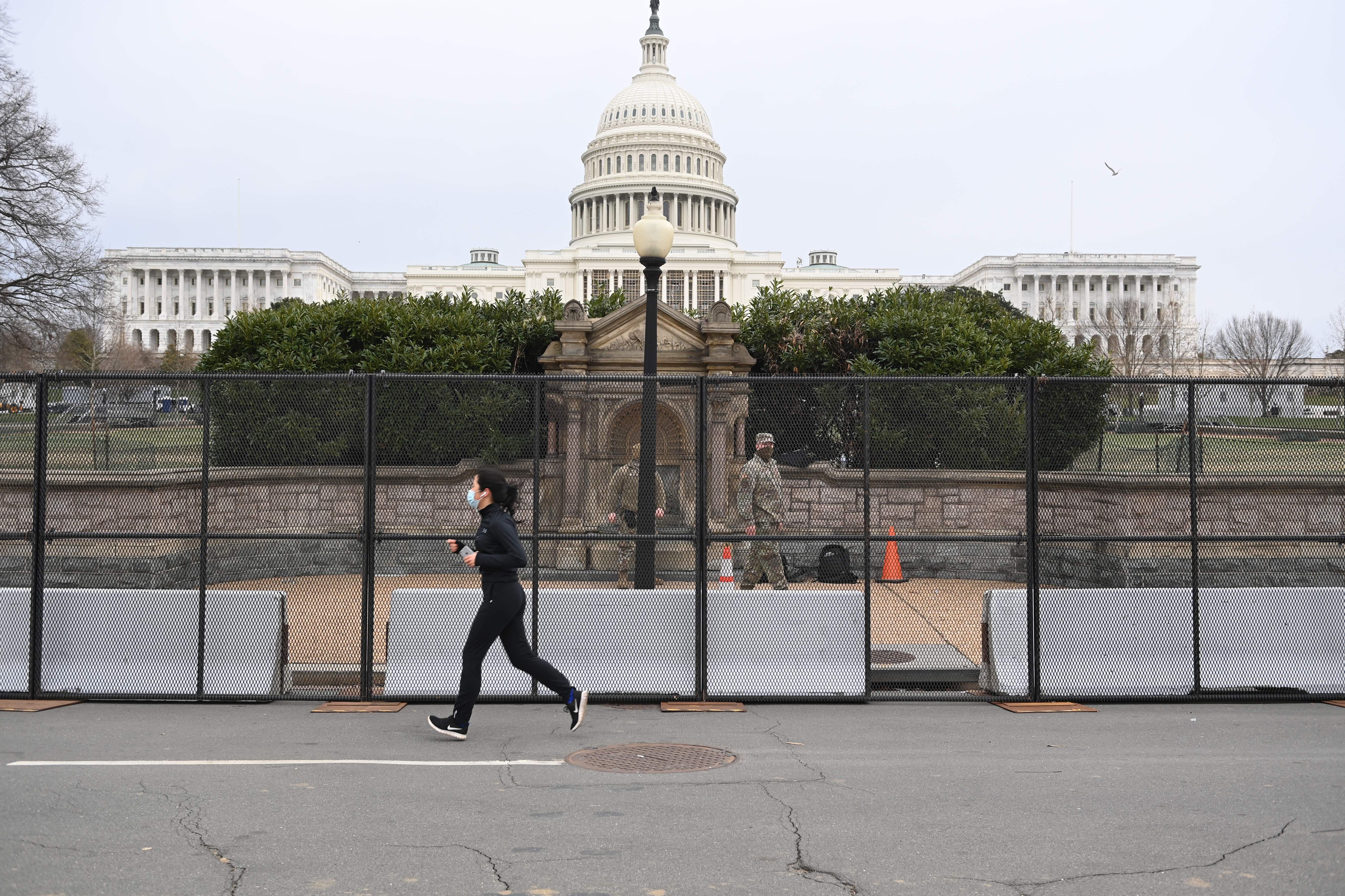 A woman jogs past a tall metal security fence in front of the low stone wall surrounding the Capitol. National Guardsmen in camo patrol the sidewalk between the fence and the stone wall, and the Capitol dome looms in the background.