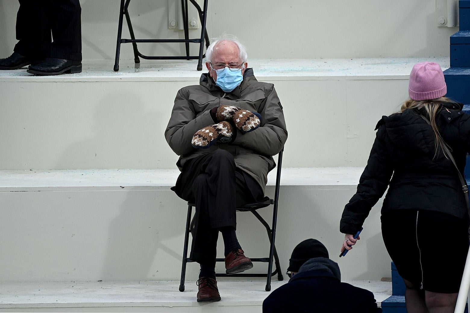 Bernie Sanders sits in a folding chair in an army green jacket and multicolored mittens with his forearms crossed.