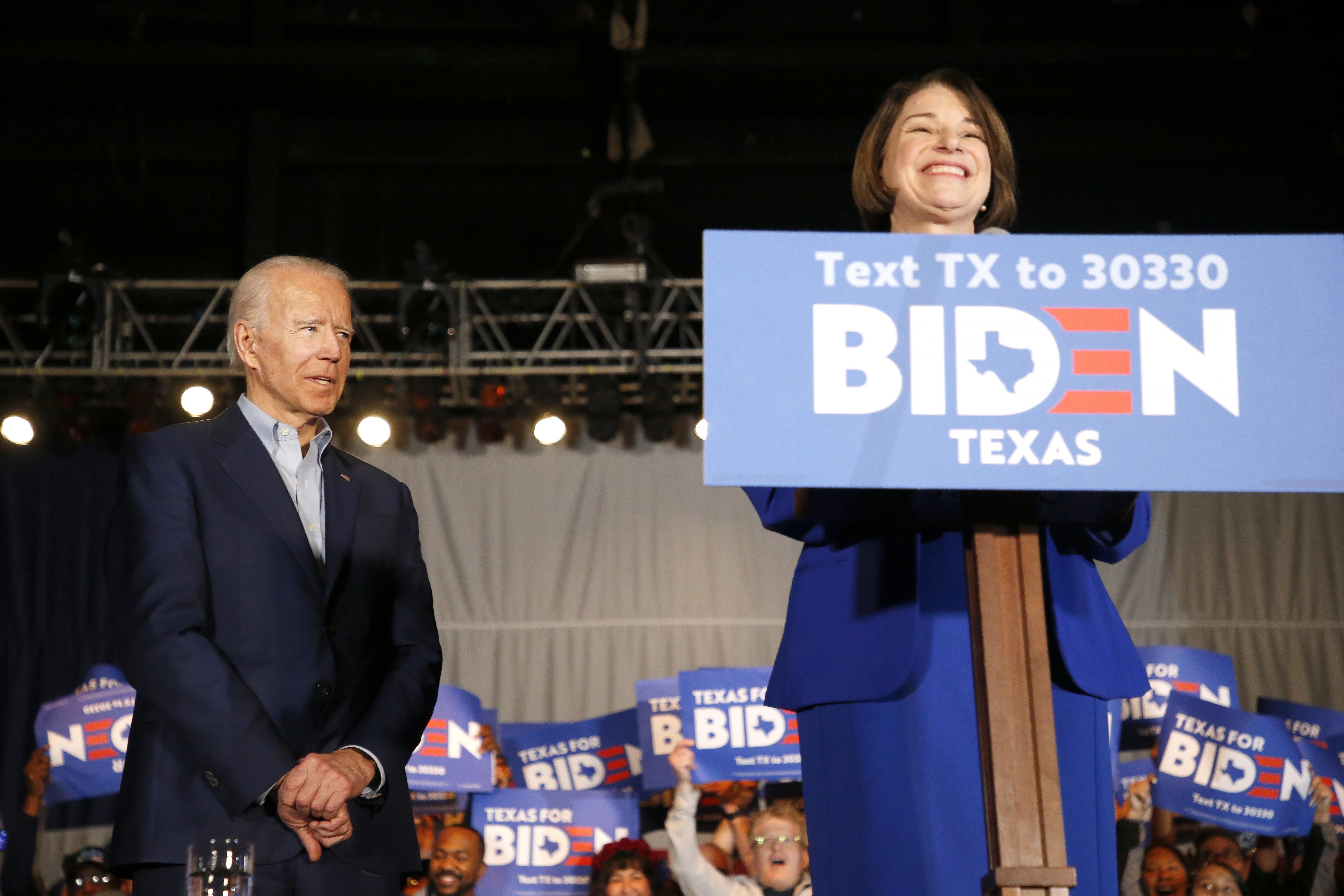 Klobuchar stands at the podium, smiling, as Biden looks on, with his supporters in the background.