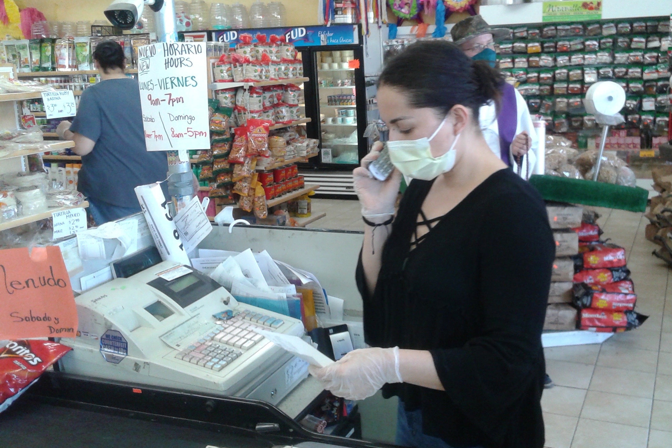 A woman wearing a mask stands behind a cash register and holds a cellphone up to her ear.