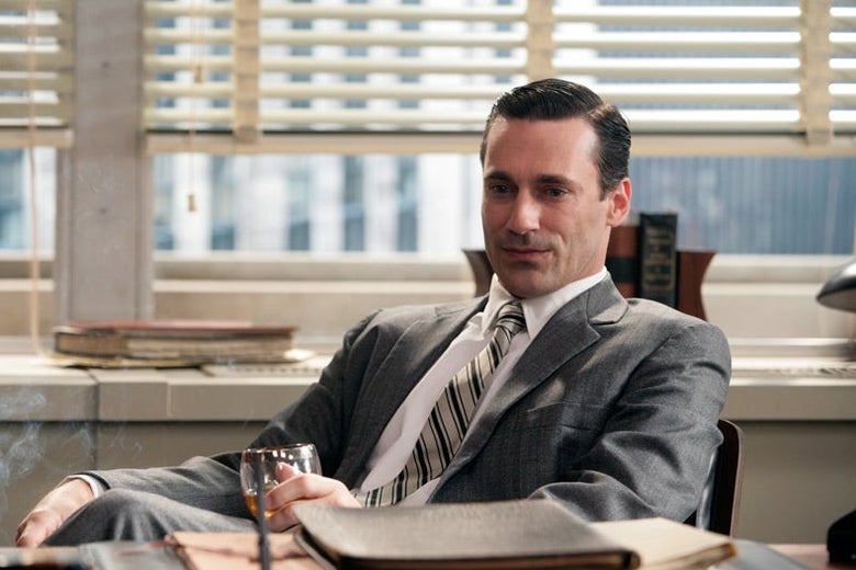 Jon Hamm in a grey suit sits behind a desk, a glass of brandy in hand.