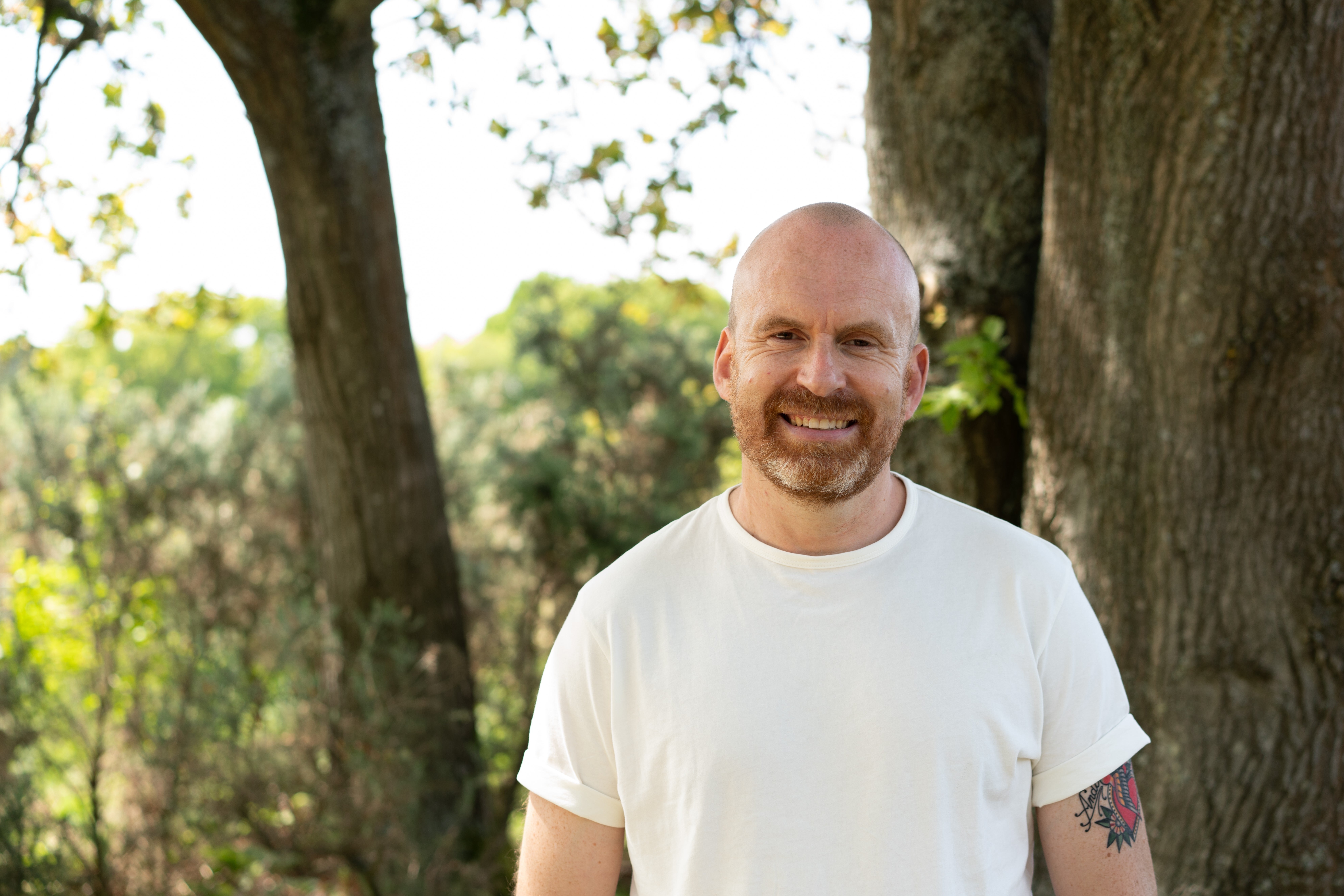 A white bald man in a white T-shirt smiles, standing in front of the trunk of a large tree.
