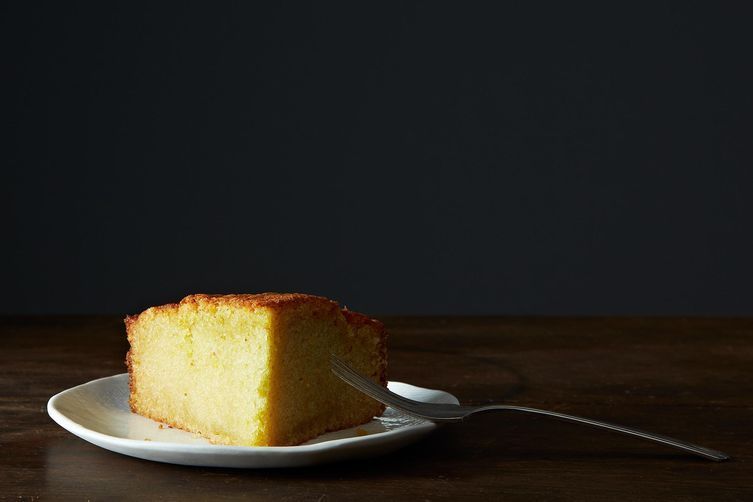 A slice of olive oil cake sits on a round, white plate on a wooden surface. A silver fork leans against the plate. The wedge side of the cake slice faces the camera, so you can see its yellow-golden inside.