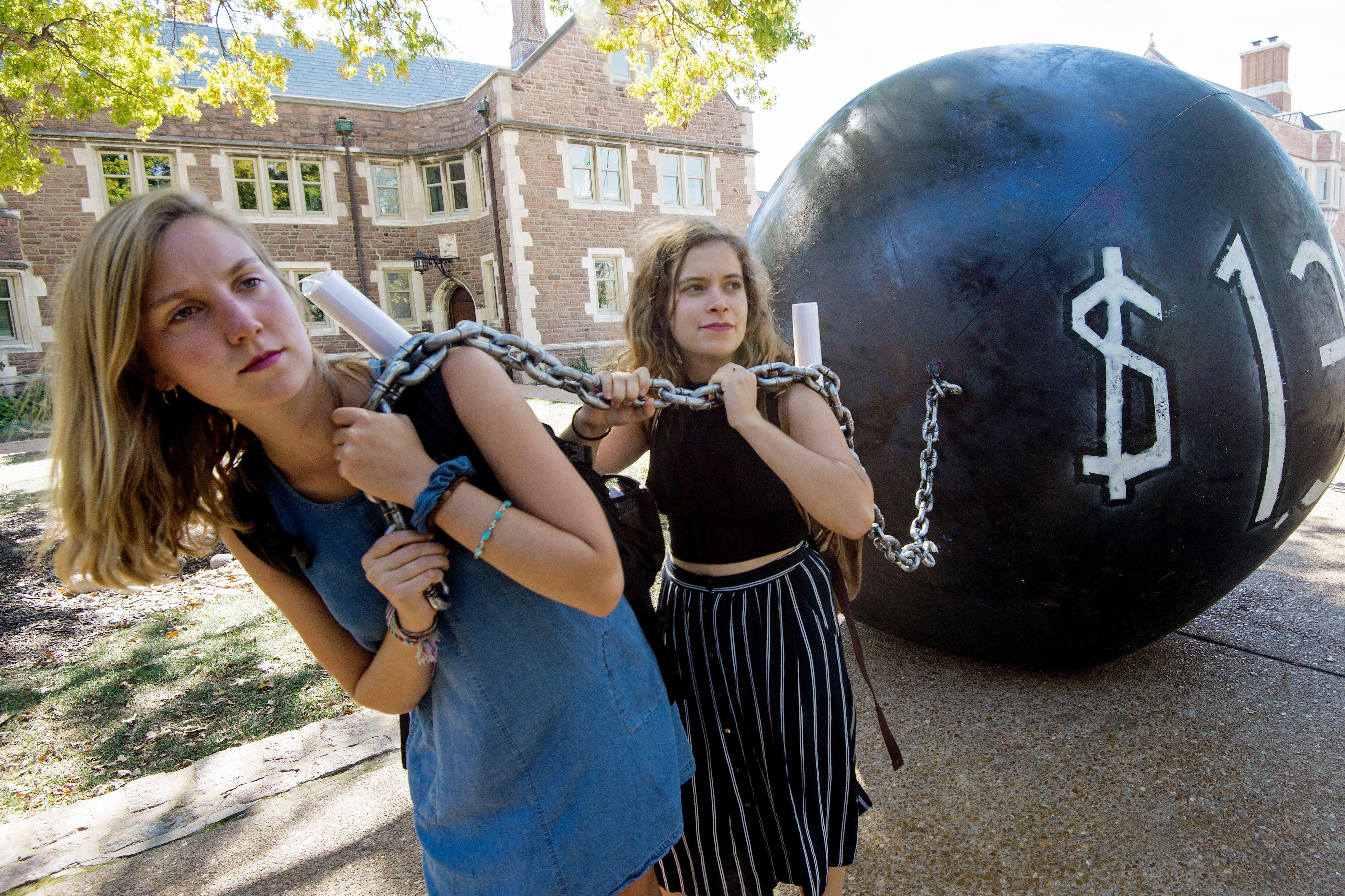 Two young women hold a chain connected to a gigantic black ball with $1.4 trillion written on it.