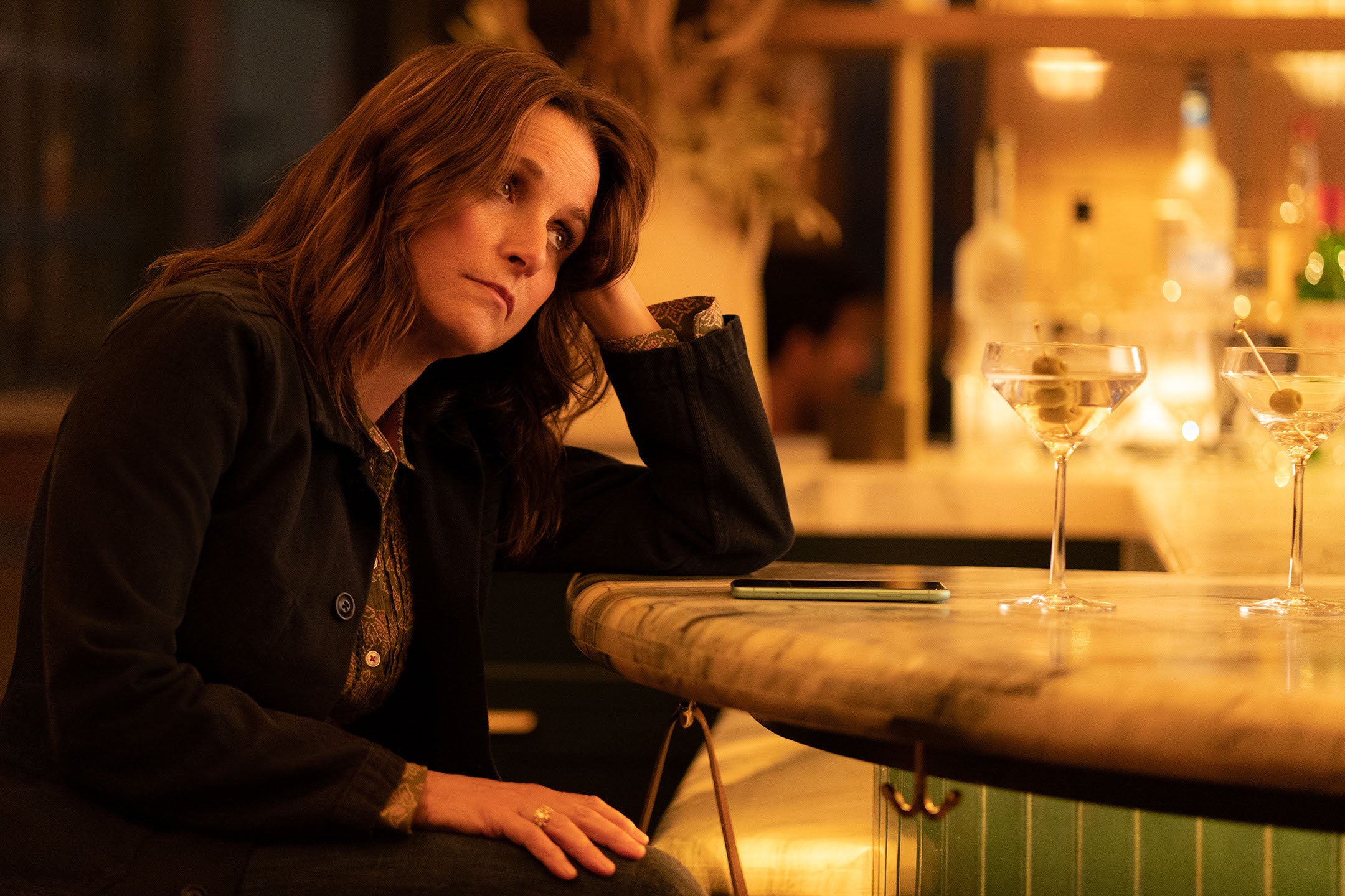 She sits at a fancy marble bar, a martini in front of her, looking somewhat despondent with her head in her left hand.