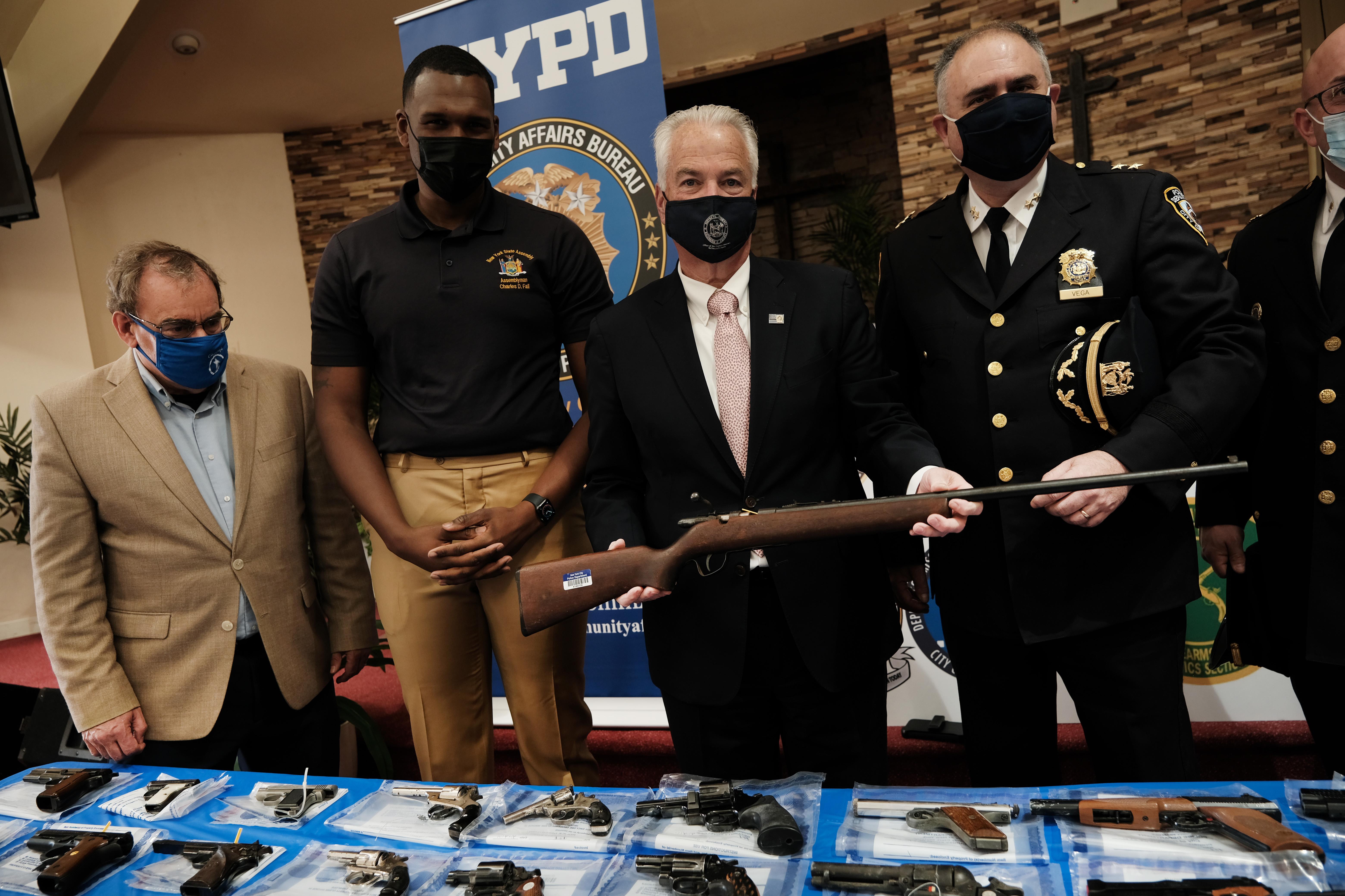 Law enforcement officials hold a rifle and stand over a table of handguns.