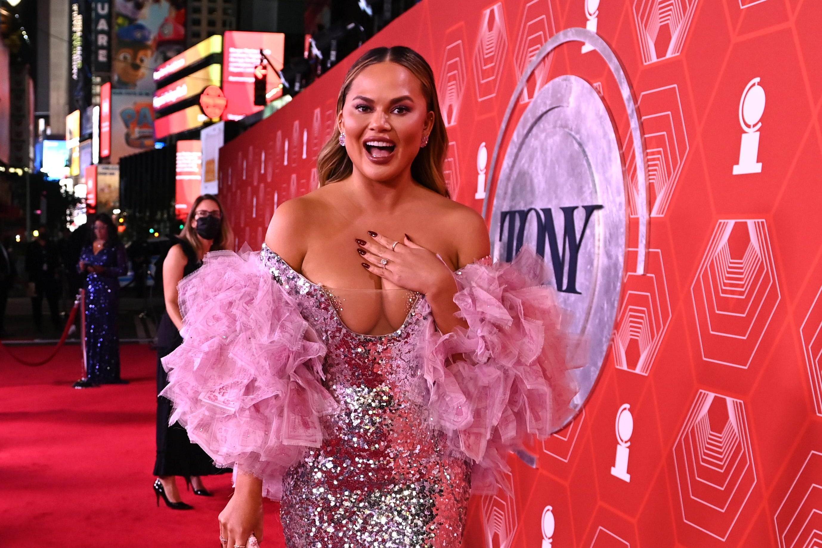 Chrissy Teigen on the red carpet in an off the shoulder silver sequin gown with pink ruffled sleeves. She is smiling and putting her left hand on her sternum as if to say "who, me?"