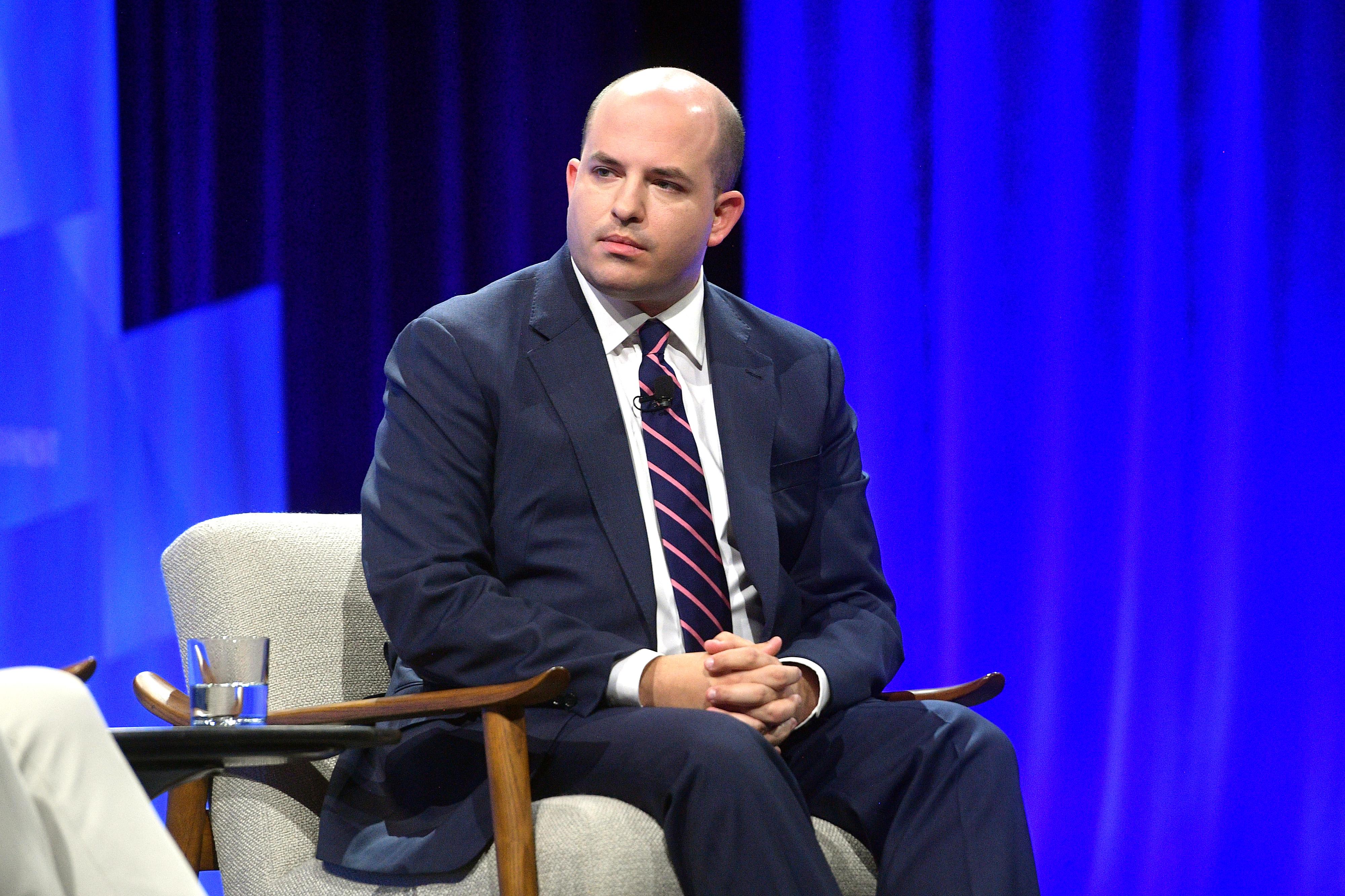Brian Stelter sitting on a chair onstage next to a table with a glass of water