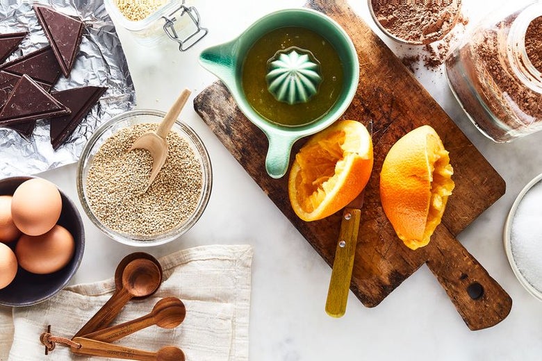 Ingredients and kitchen devices are artfully scattered around a counter: chocolate, eggs, quinoa, tablespoons, a juicer, juiced oranges, a cutting board, and cocoa powder can all be seen.