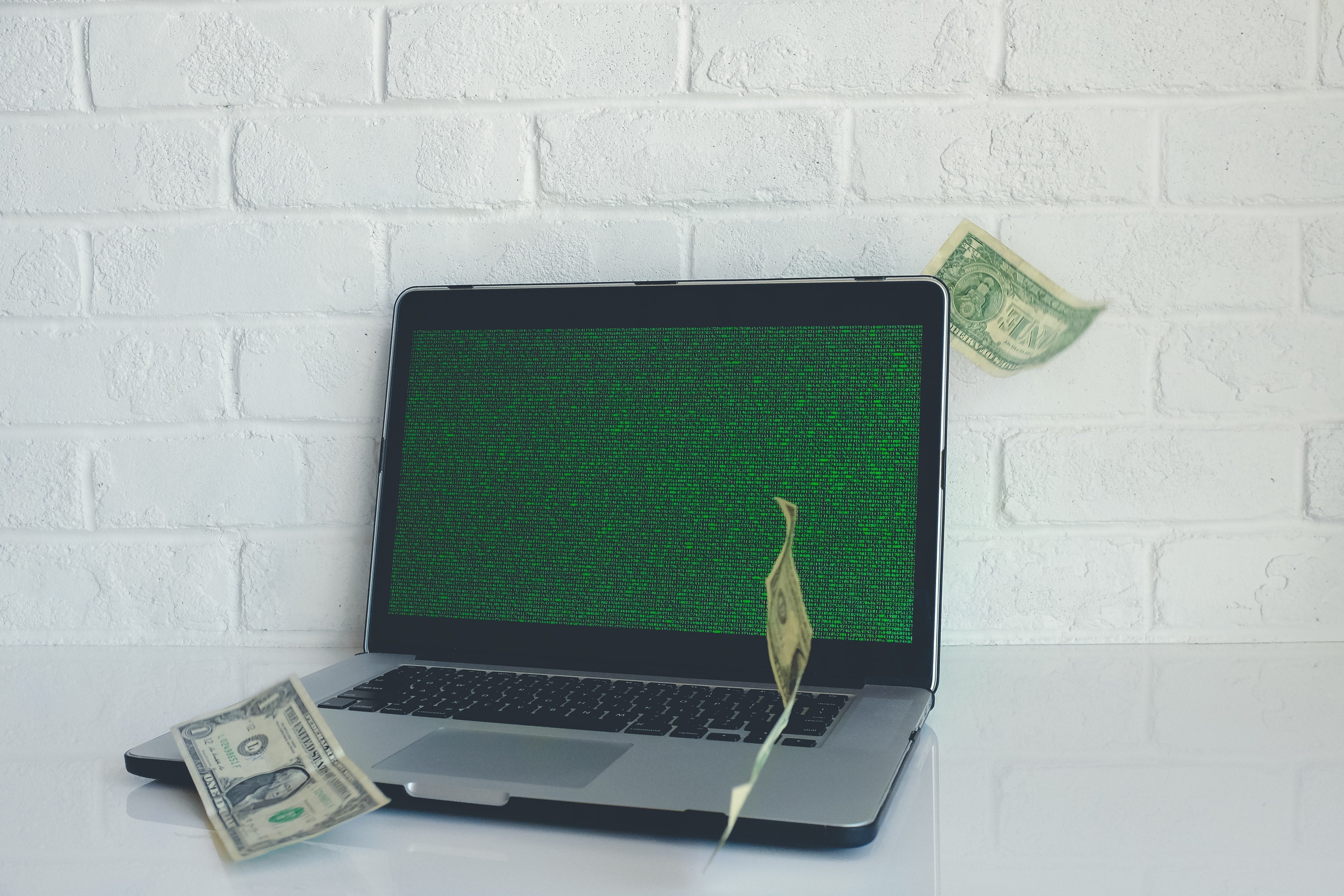 A laptop with green noise on the screen sits on a counter, with dollar bills floating around it.