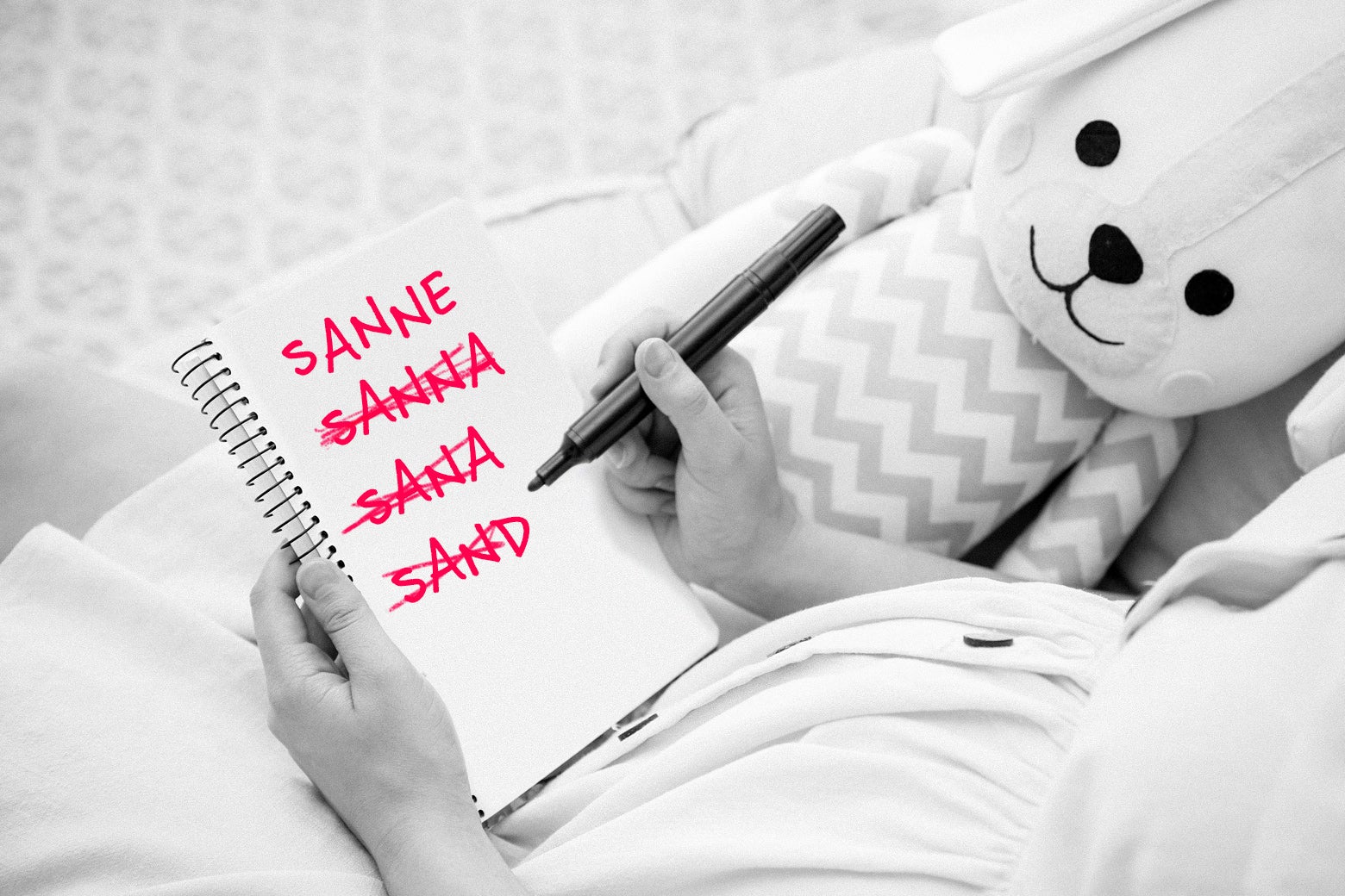 A mom holds a notebook with multiple spellings of the same name written down, next to a stuffed animal.