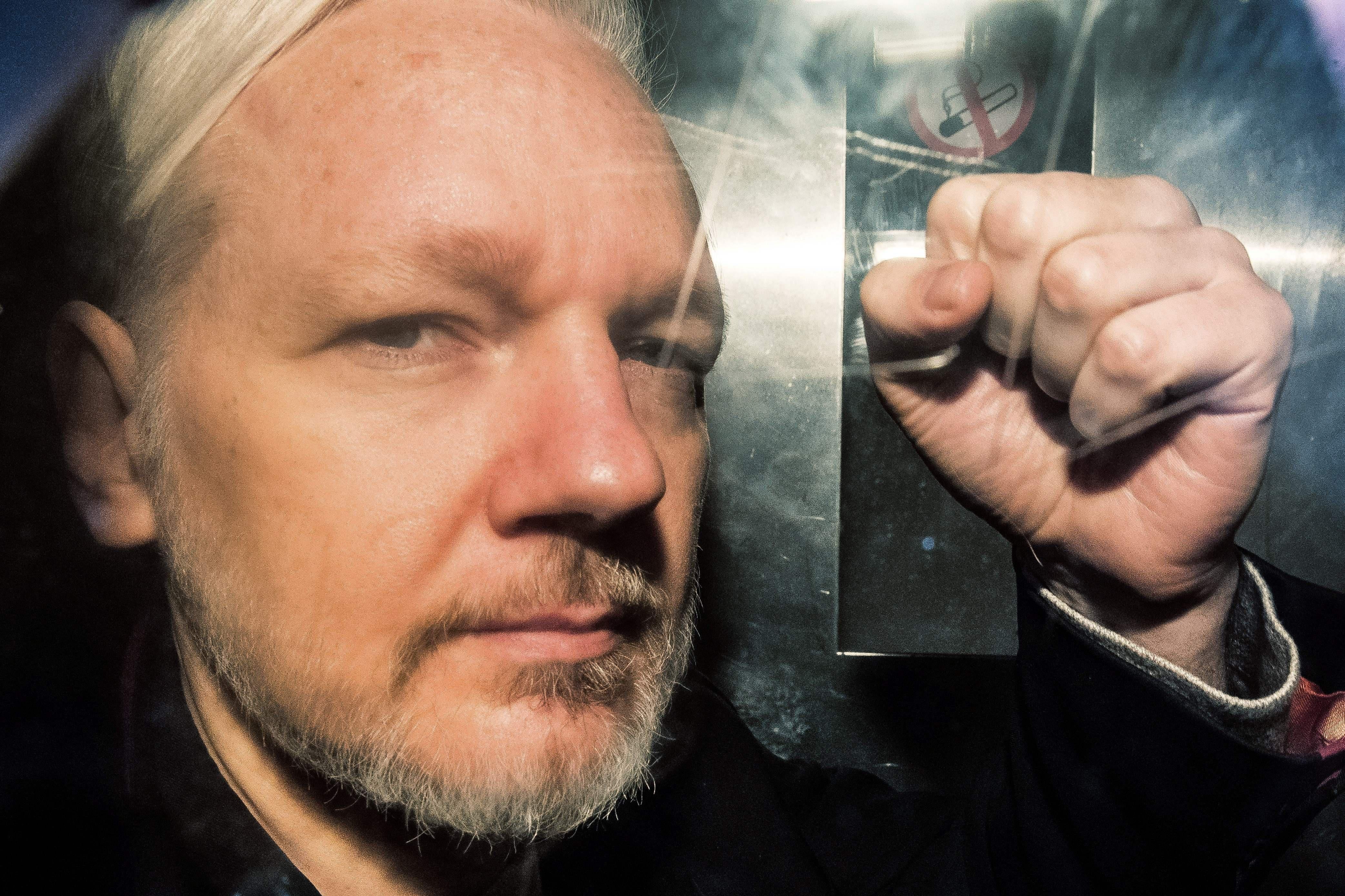 Julian Assange, seen up close through a window, gestures with a hand in a fist.