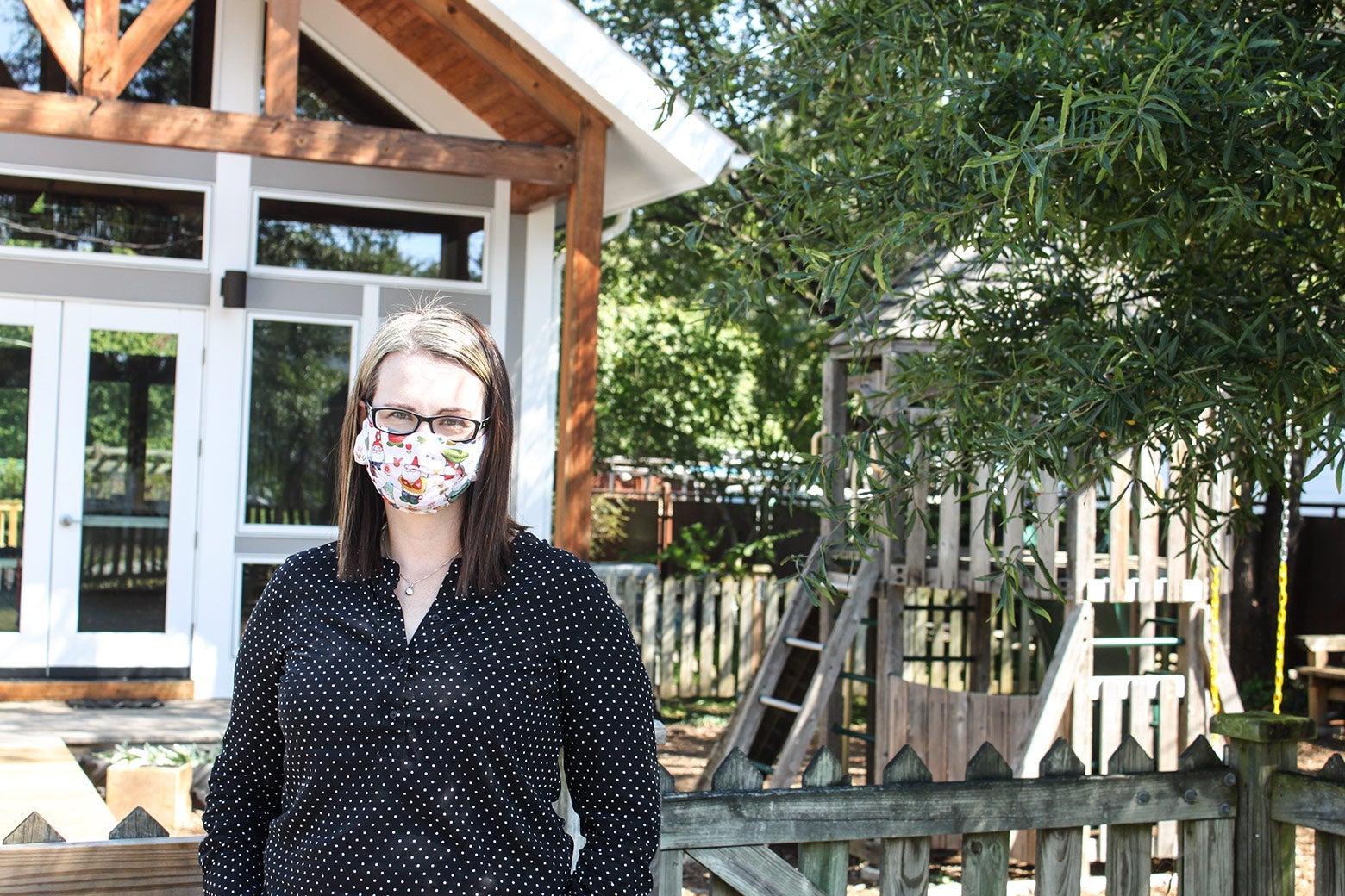 A woman in a mask stands in front of a wooden, fenced-in playground.