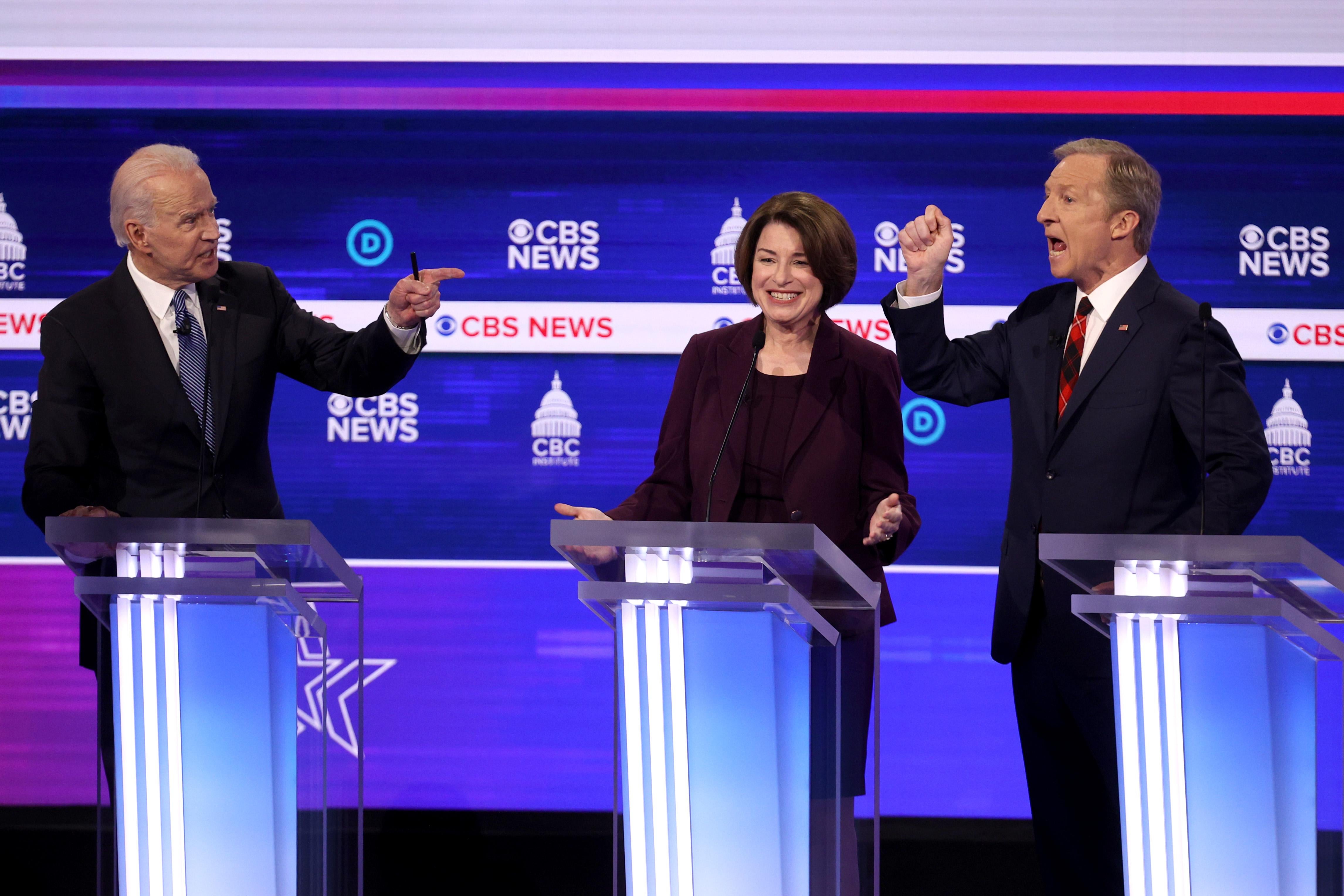 Joe Biden and Tom Steyer point angrily at each other as they speak on the debate stage. Amy Klobuchar, standing between them, smiles.