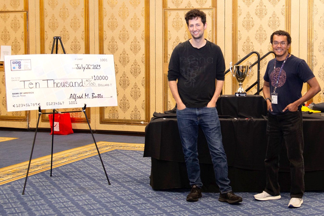 Both finalists stand looking relaxed with hands in pockets near a novelty-size $10,000 check on display on an easel. 