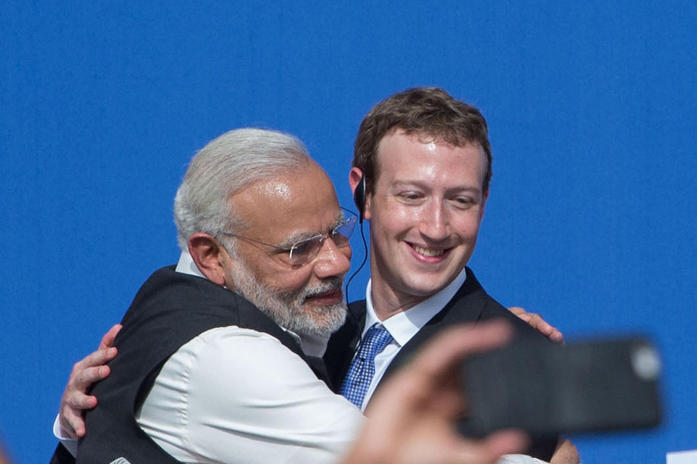 A hand holding a smartphone appears to take a photo of Modi and Zuckerberg embracing.
