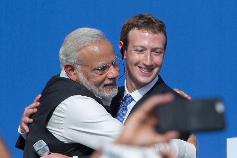 A hand holding a smartphone appears to take a photo of Modi and Zuckerberg embracing.

