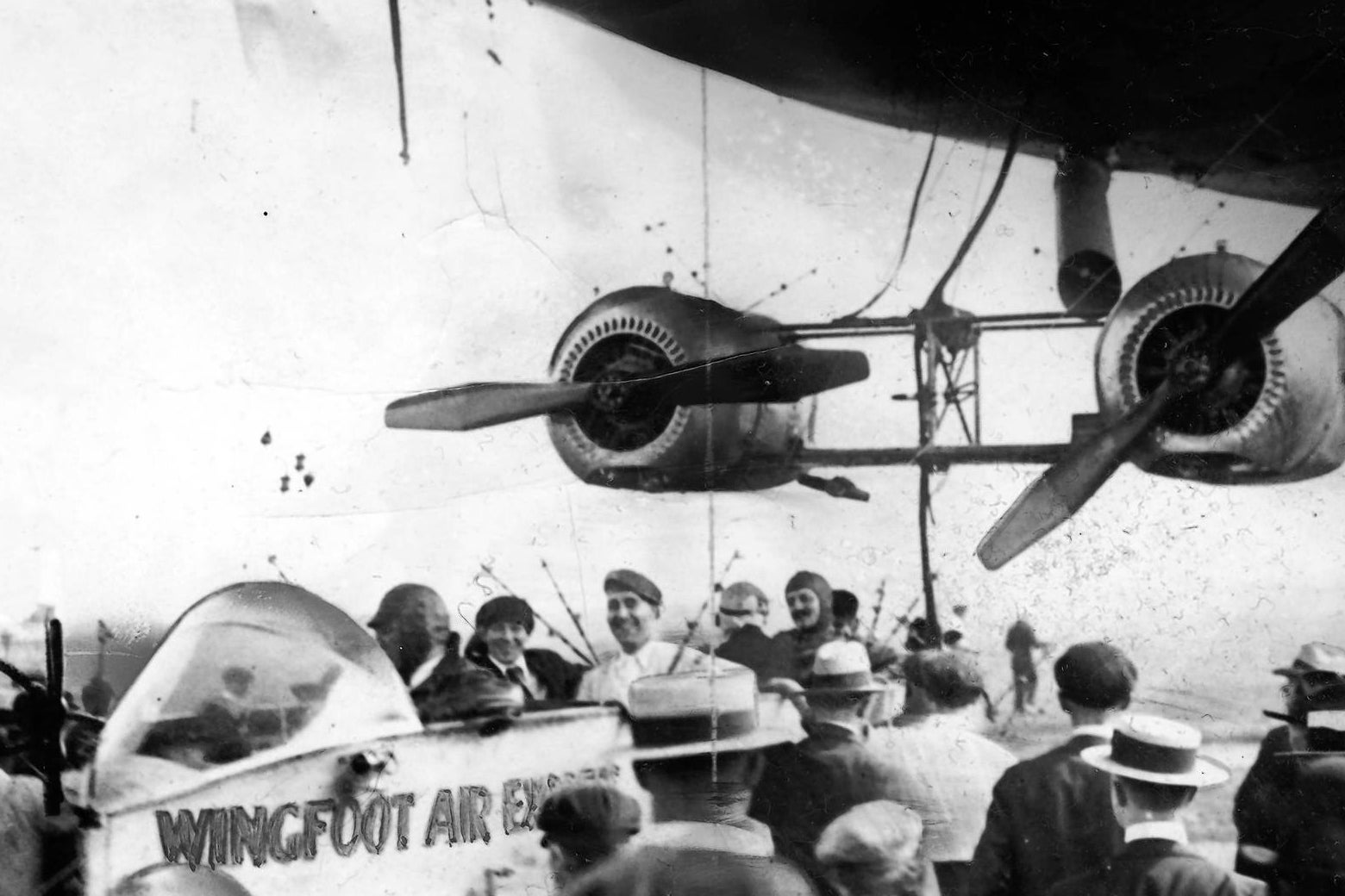 The open gondola of a 1910s blimp, flanked by two giant engines. Five smiling passengers sit in the gondola.