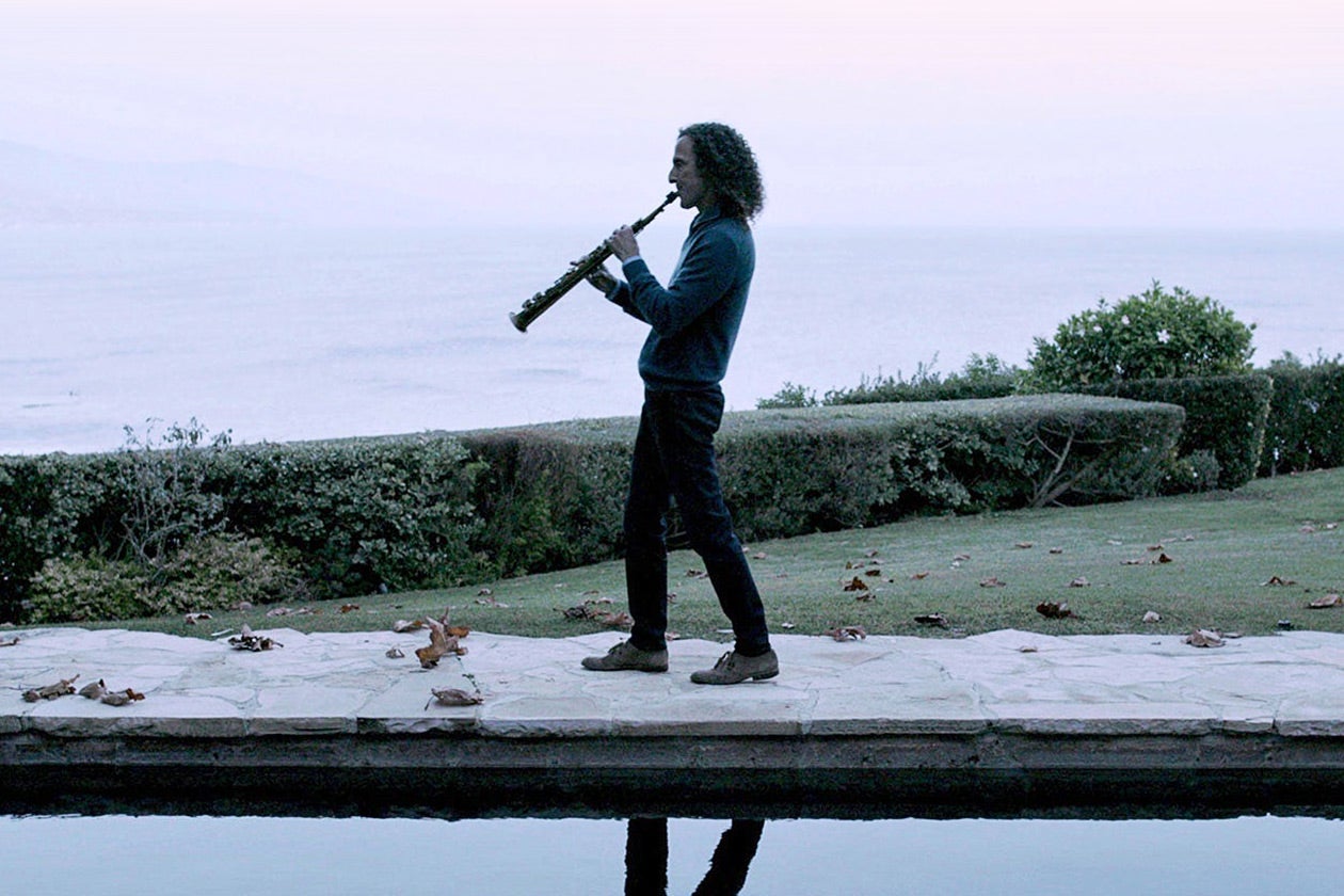 Kenny G plays his saxophone while walking by a pool showing his reflection.