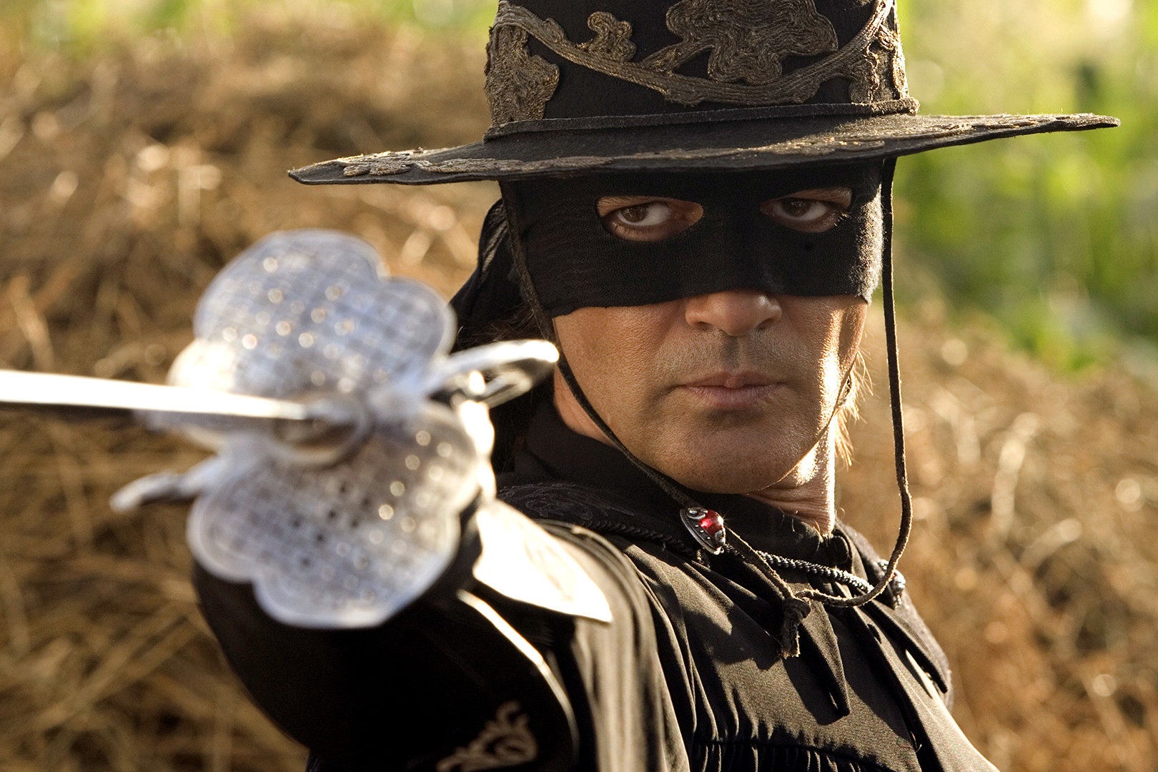 A straightfaced man dressed in a black cloak, a black mask that covers the upper half of his face (showing only his eyes and lower face), and embroidered black hat with a large brim, faces someone slightly off camera and points a silver sword in their direction. 