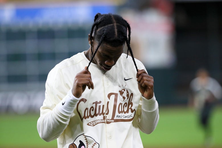 Travis Scott stands on a baseball field, holding his braids. His shirt says Cactus Jack Foundation. 