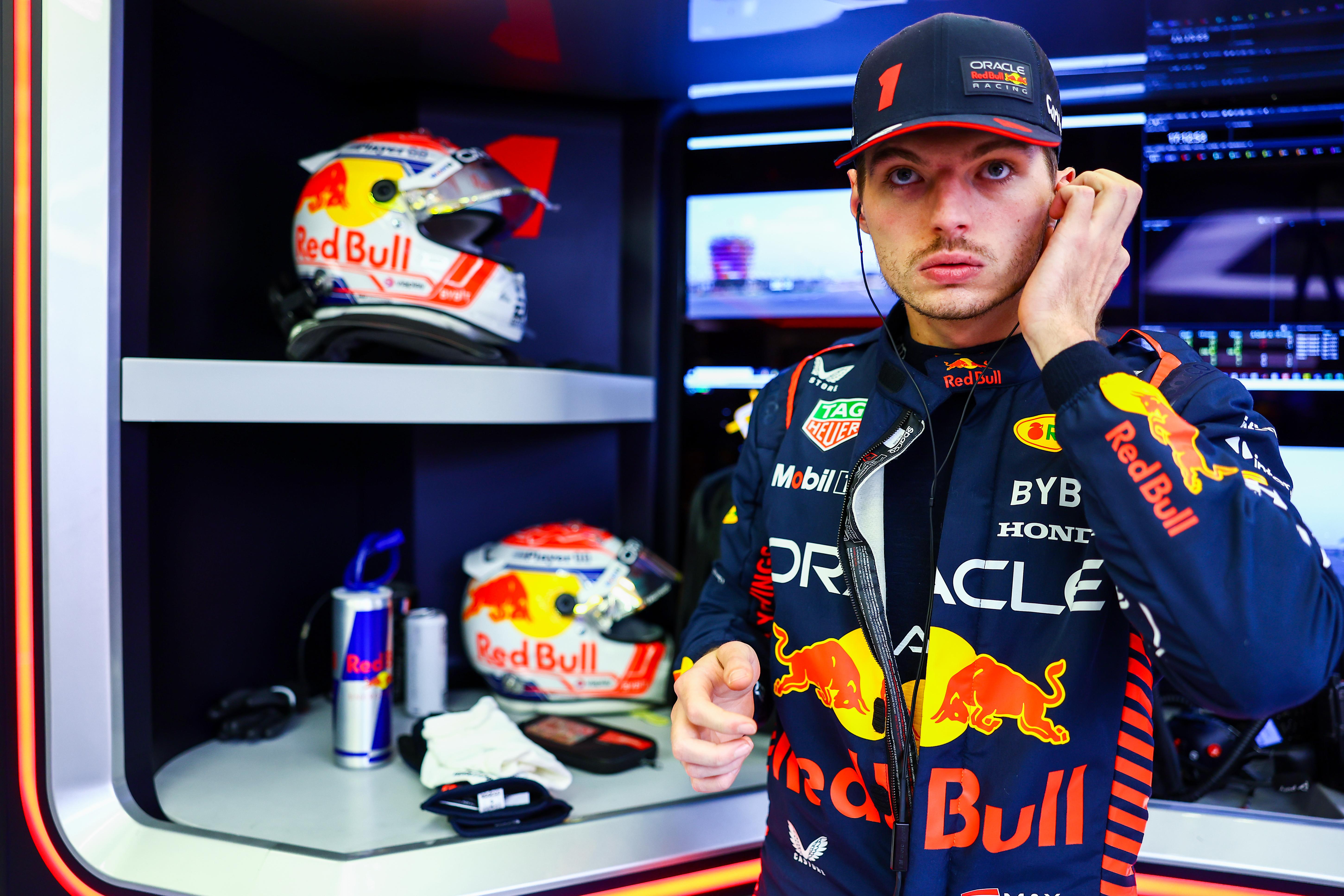 Verstappen surrounded by Red Bull logo'd gear in his racing uniform, looking stern-faced with his left hand to his ear