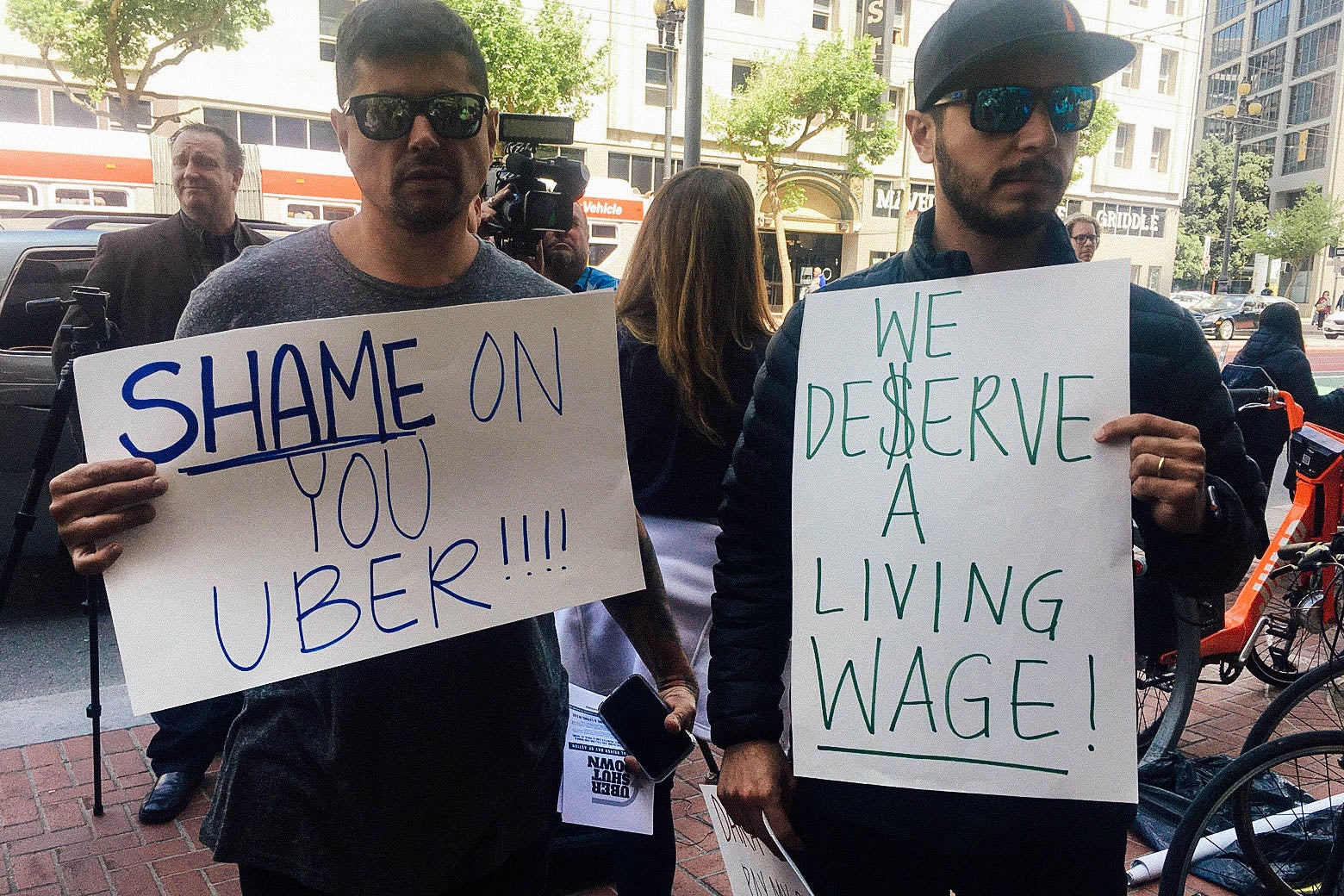 Protesters holding signs reading "SHAME ON YOU UBER" AND "WE DESERVE A LIVING WAGE."