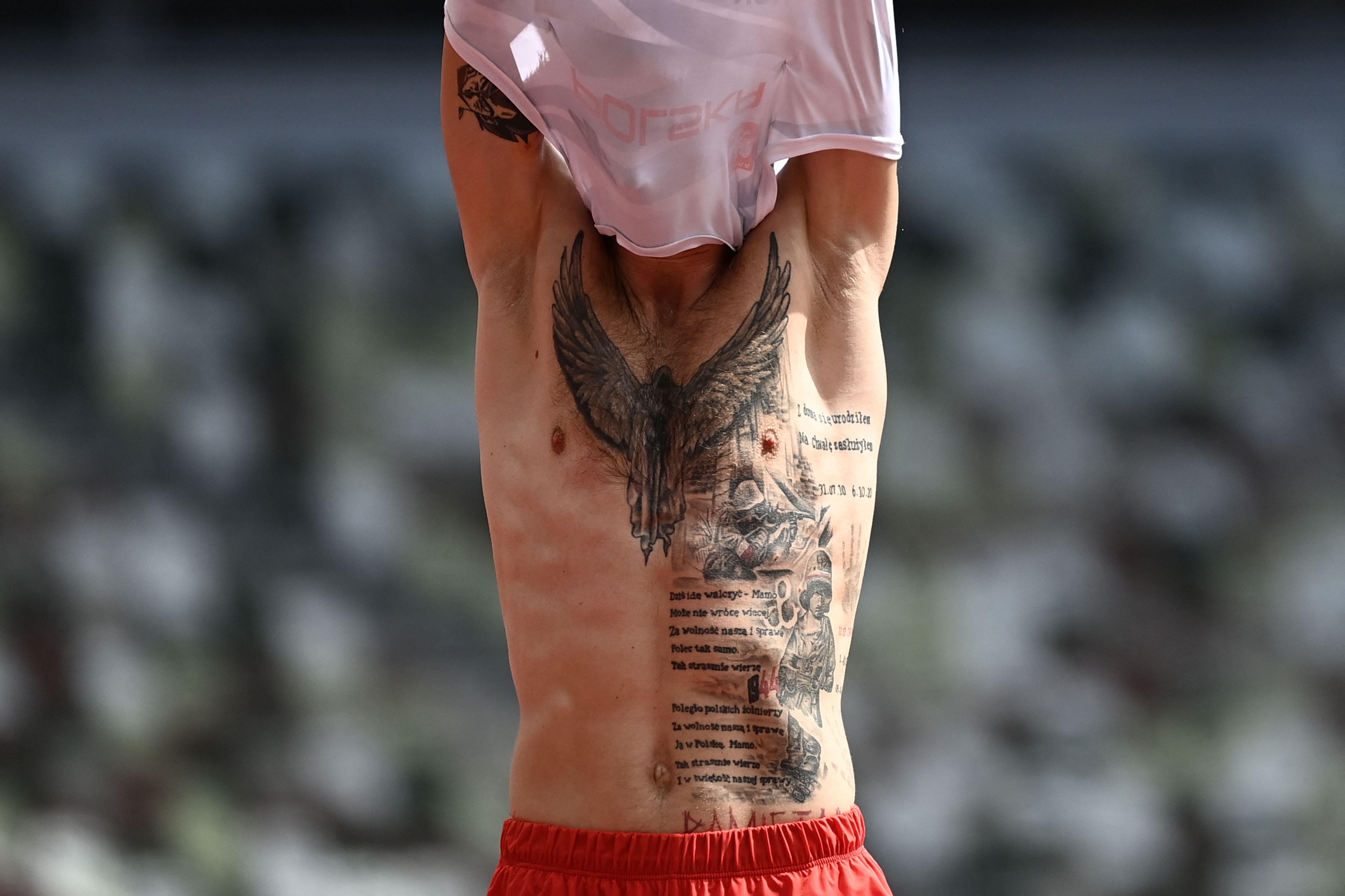 A man lifts his shirt over his head revealing extensive tattoos on his chest and abdomen, including a large bird on his chest and ample text on his stomach.