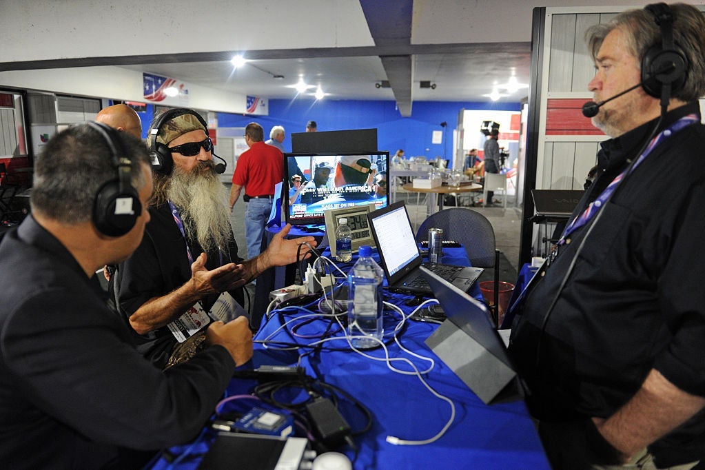 Bossie, a broad-shouldered man with short graying black hair, sits at a table in a bustling media room with the long-bearded Phil Robertson of Duck Dynasty. Steve Bannon is standing next to the table. All three are wearing headsets for a radio broadcast.