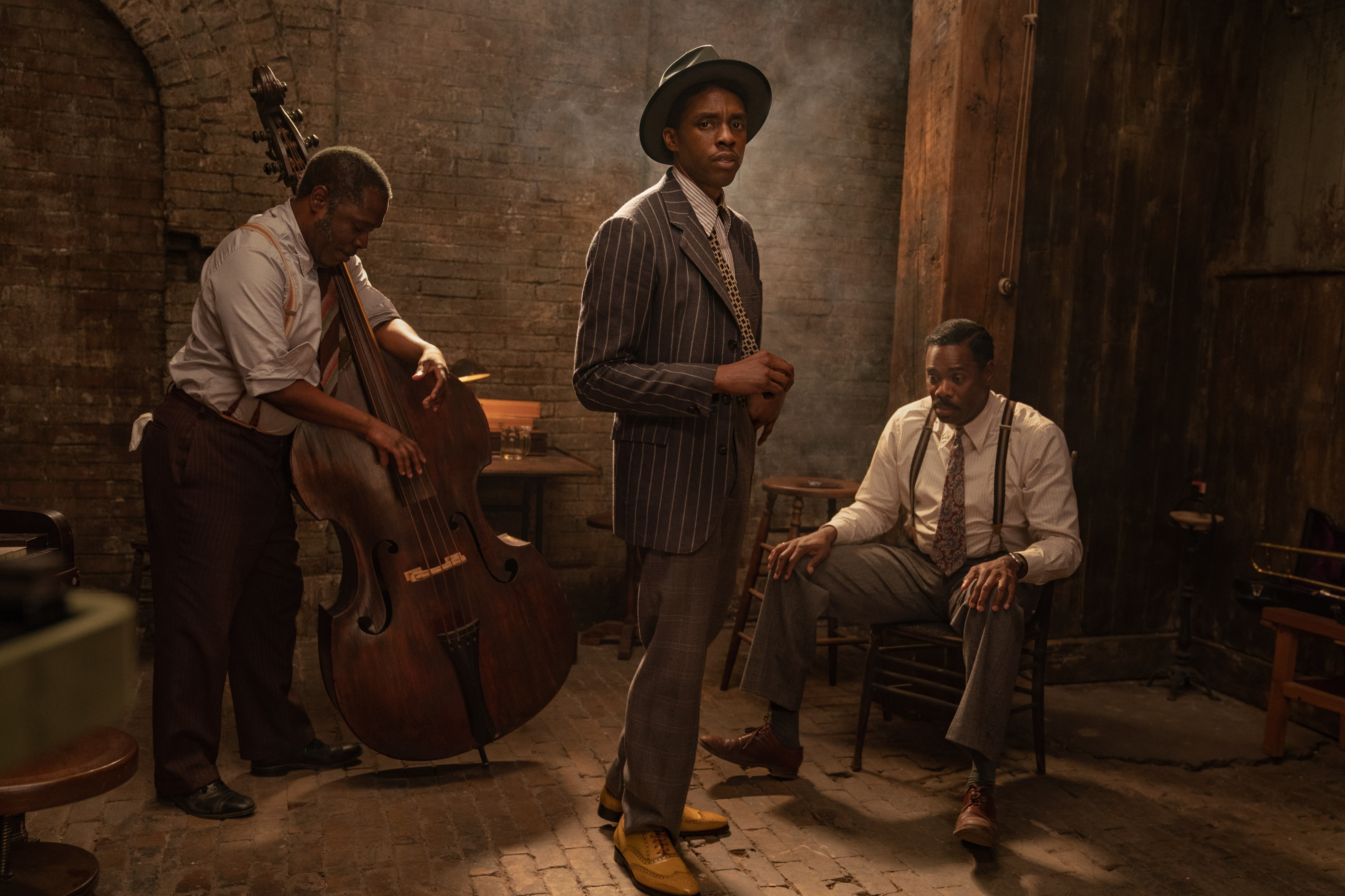 Chadwick Boseman stands in a pinstriped suit and fedora, in front of a man with an upright bass and another man seated in a chair.