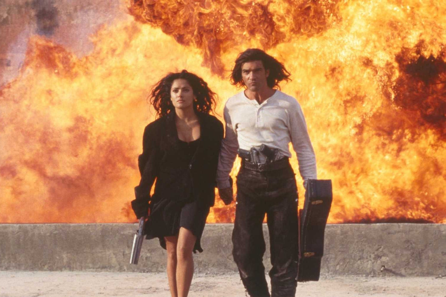 Salma Hayek (left) and Antonio Banderas (right) hold hands and a gun and guitar case (respectively) as they walk away straight faced as an explosion unfolds behind them.