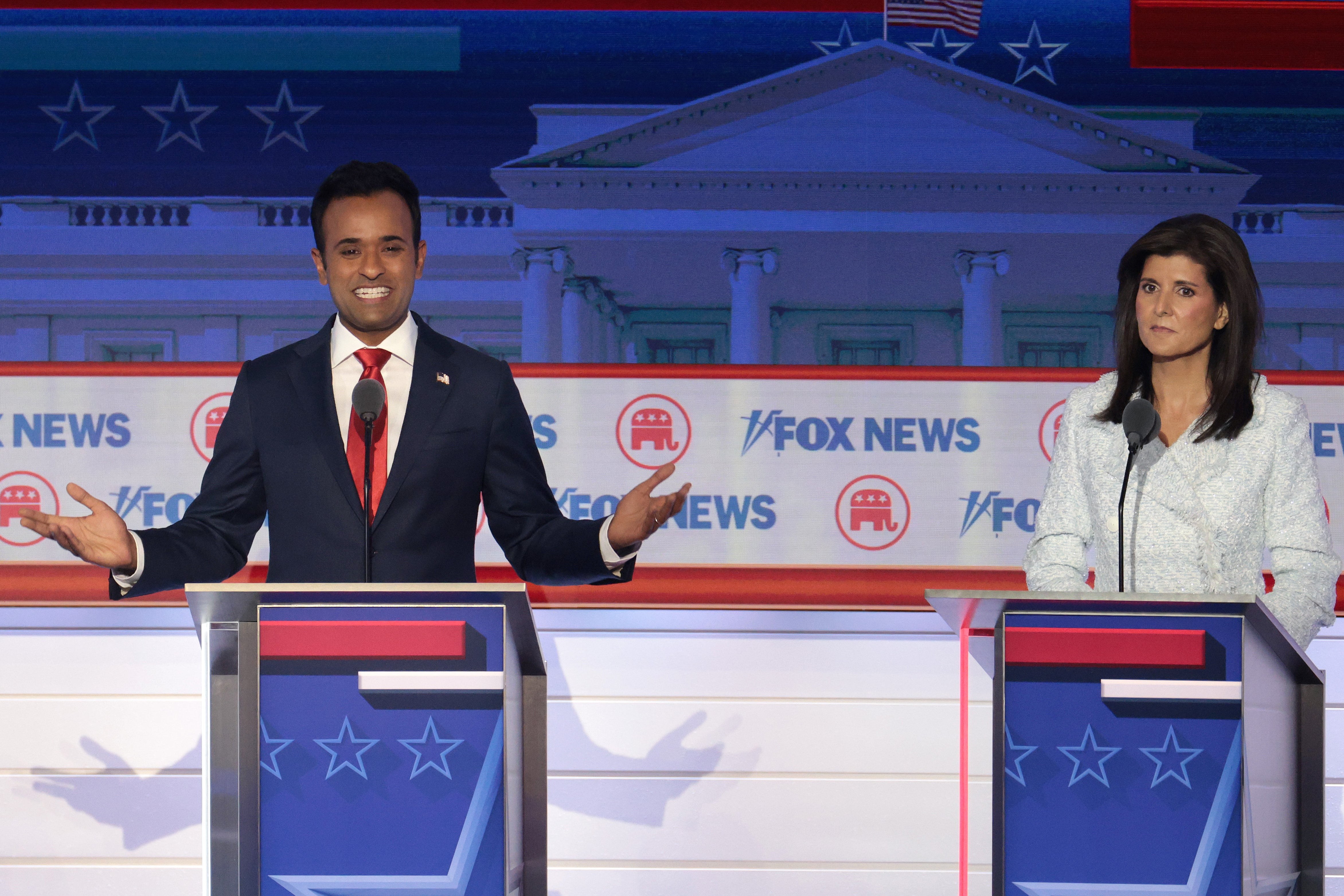 A grinning Ramaswamy holds his palms up to his side at his debate lectern as Nikki Haley, to his right, wears an exhausted expression.