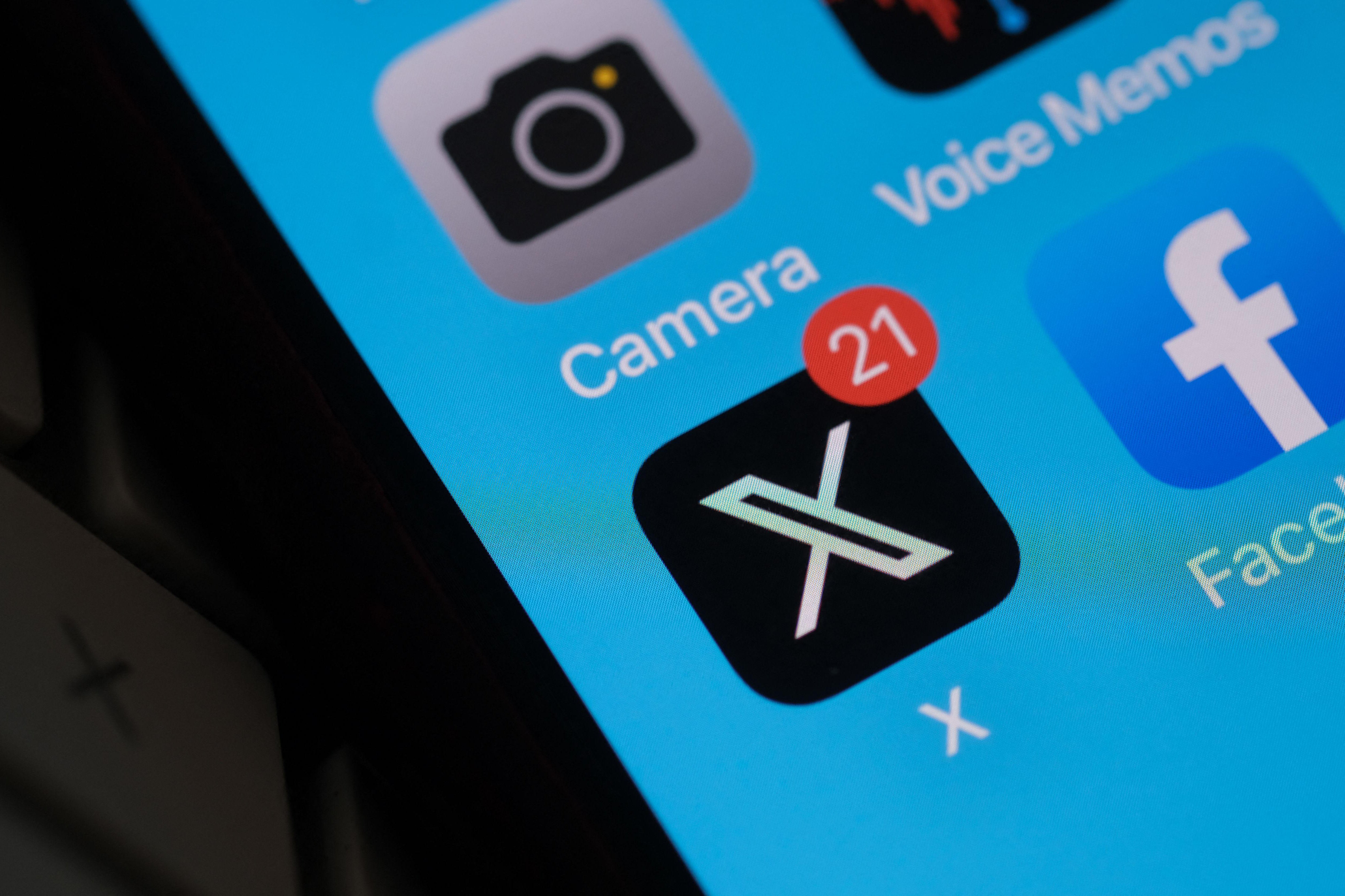 The X app's icon, with a badge displaying the presence of 21 notifications, is seen on an iPhone screen under the Camera app and next to the Facebook app.