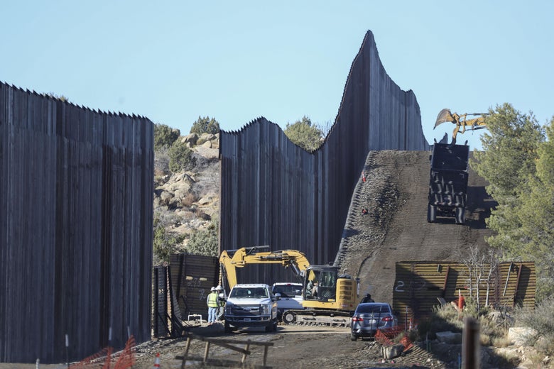 Construction vehicles and workers next to a section of tall fencing with a gap in it