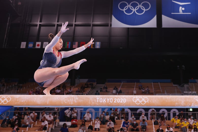 Lee mid–wolf turn on balance beam, left leg crouched on the beam, right outstretched, arms raised into the air