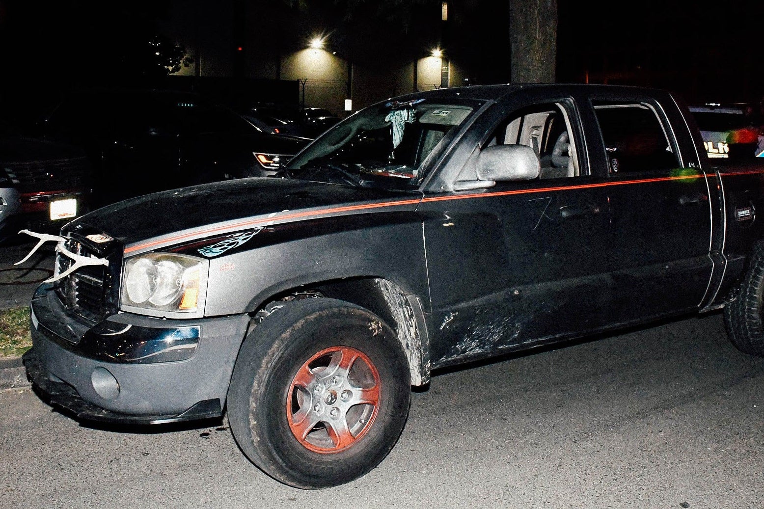 A dilapidated black truck with red piping and what appear to be antlers on its front grille is seen in the glare of a camera flash or spotlight.