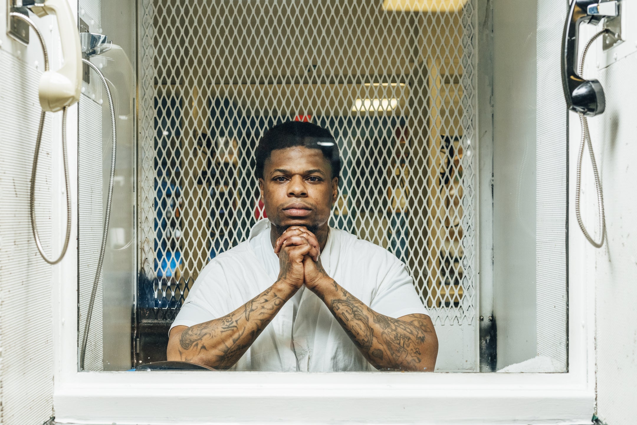 A man sits in a prison visitation booth with his hands clasped together as he looks toward the camera