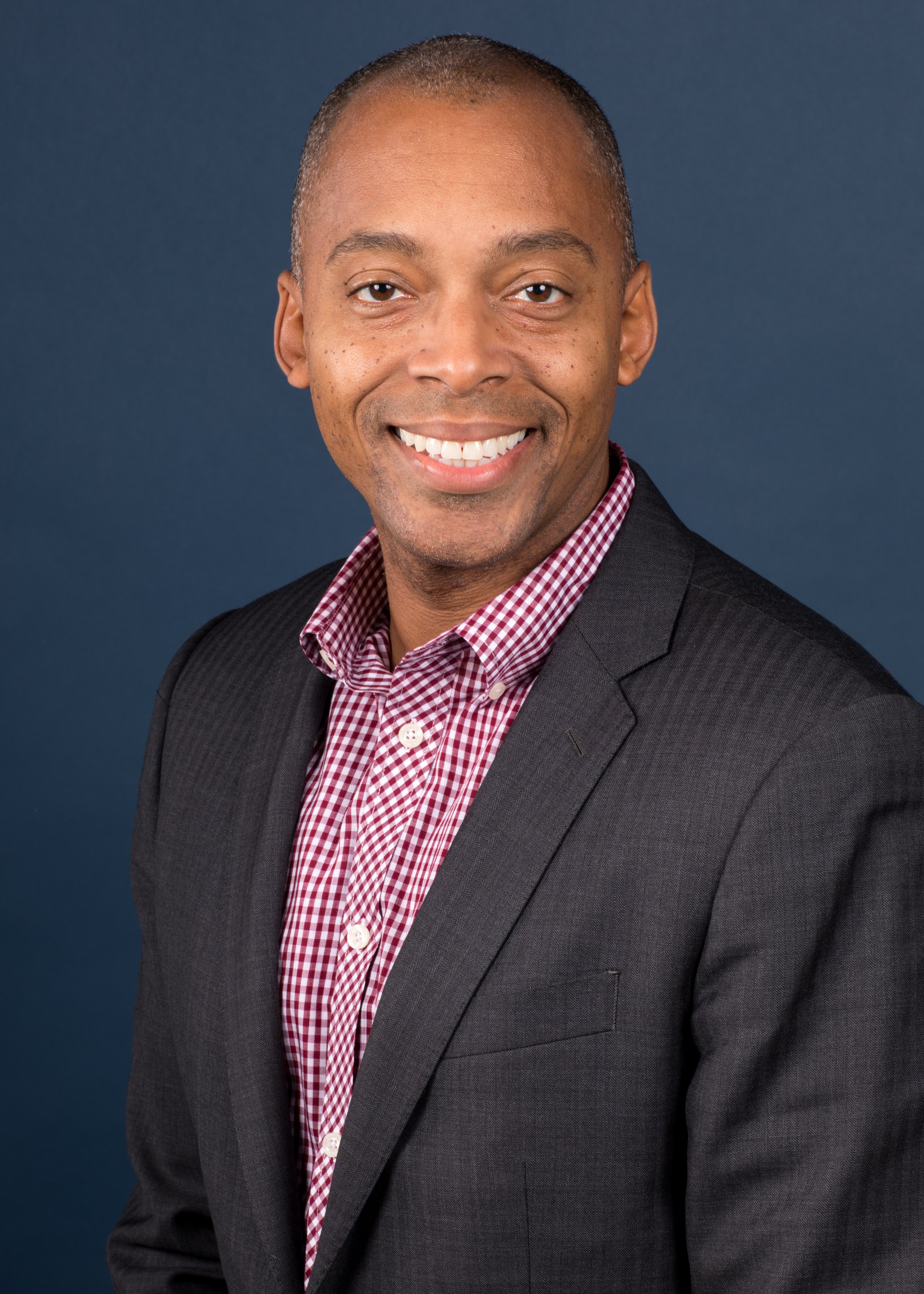 A smiling Black man in a checked shirt and blazer.