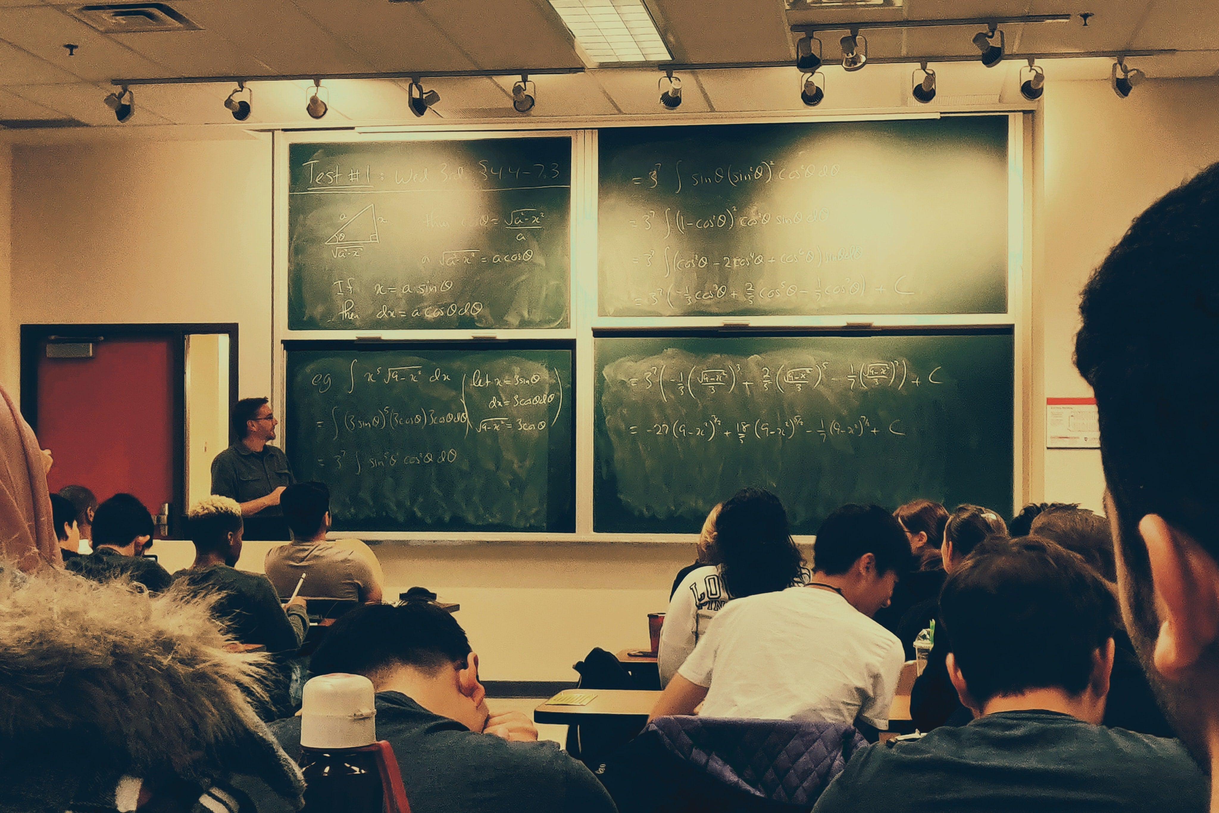 A professor stands in front of a chalkboard and lectures to students sitting at desks in a classroom