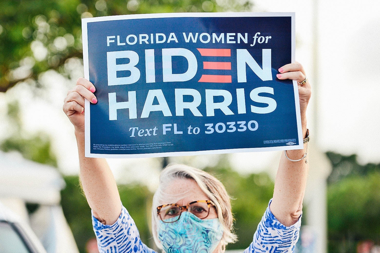 A white woman in glasses and a face mask holds up a sign that says, "Florida women for Biden Harris."