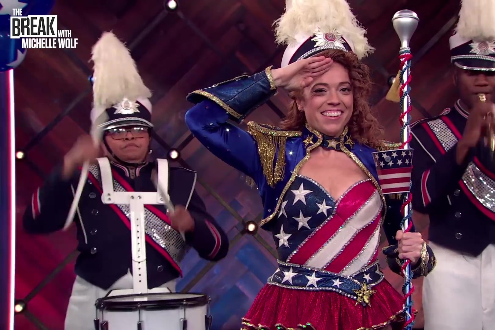 Michelle Wolf, dressed in red, white, and blue, salutes abortion as a marching band plays behind her.