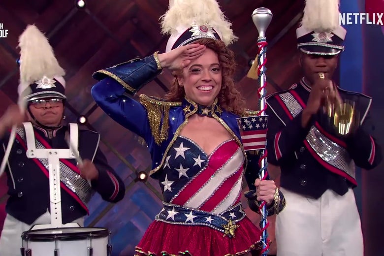 Michelle Wolf, dressed in red, white, and blue, salutes abortion as a marching band plays behind her.