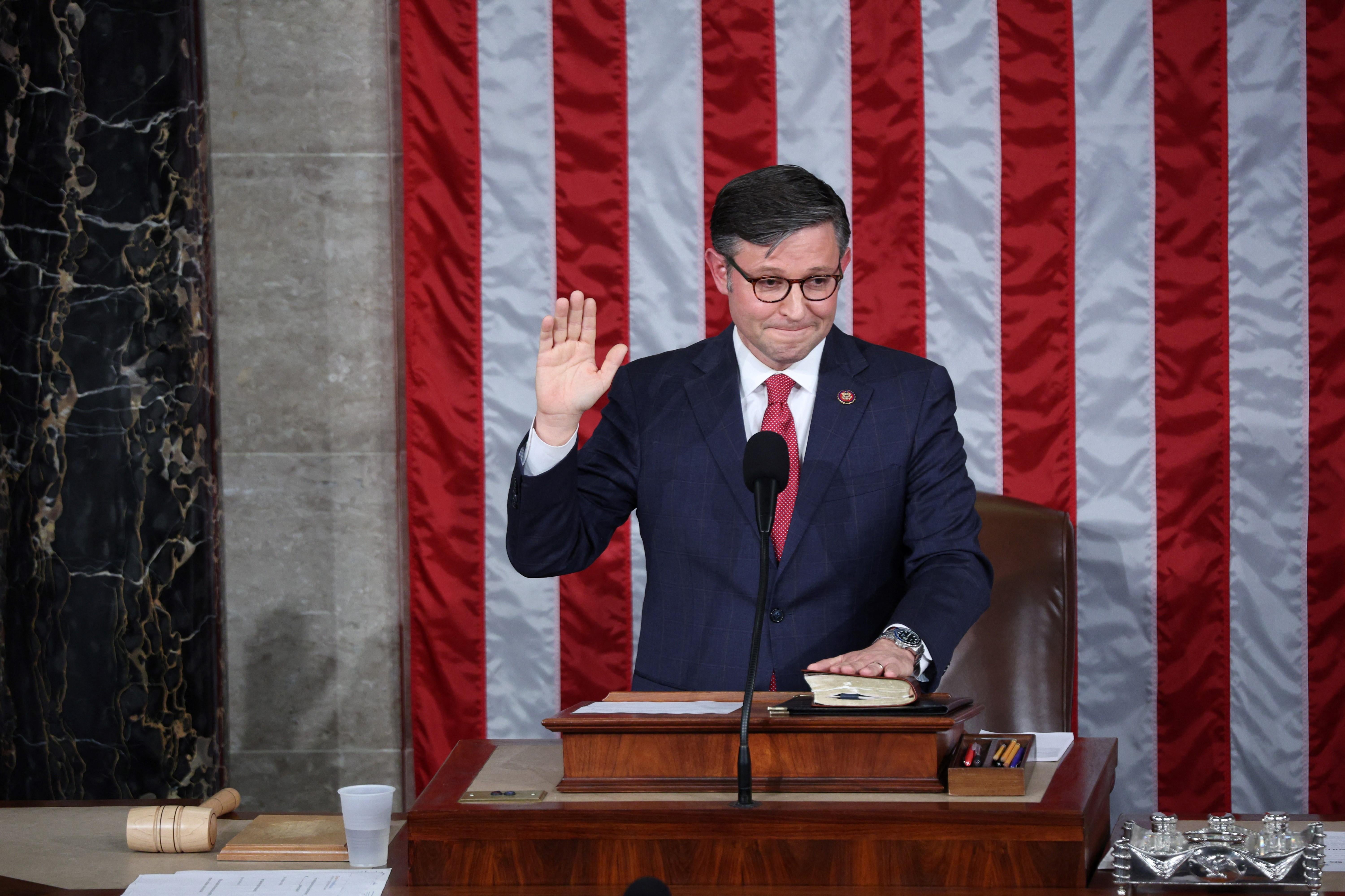Mike Johnson places one hand on a book and raises his other hand while standing in front of a flag on the dais in the U.S. Capitol.