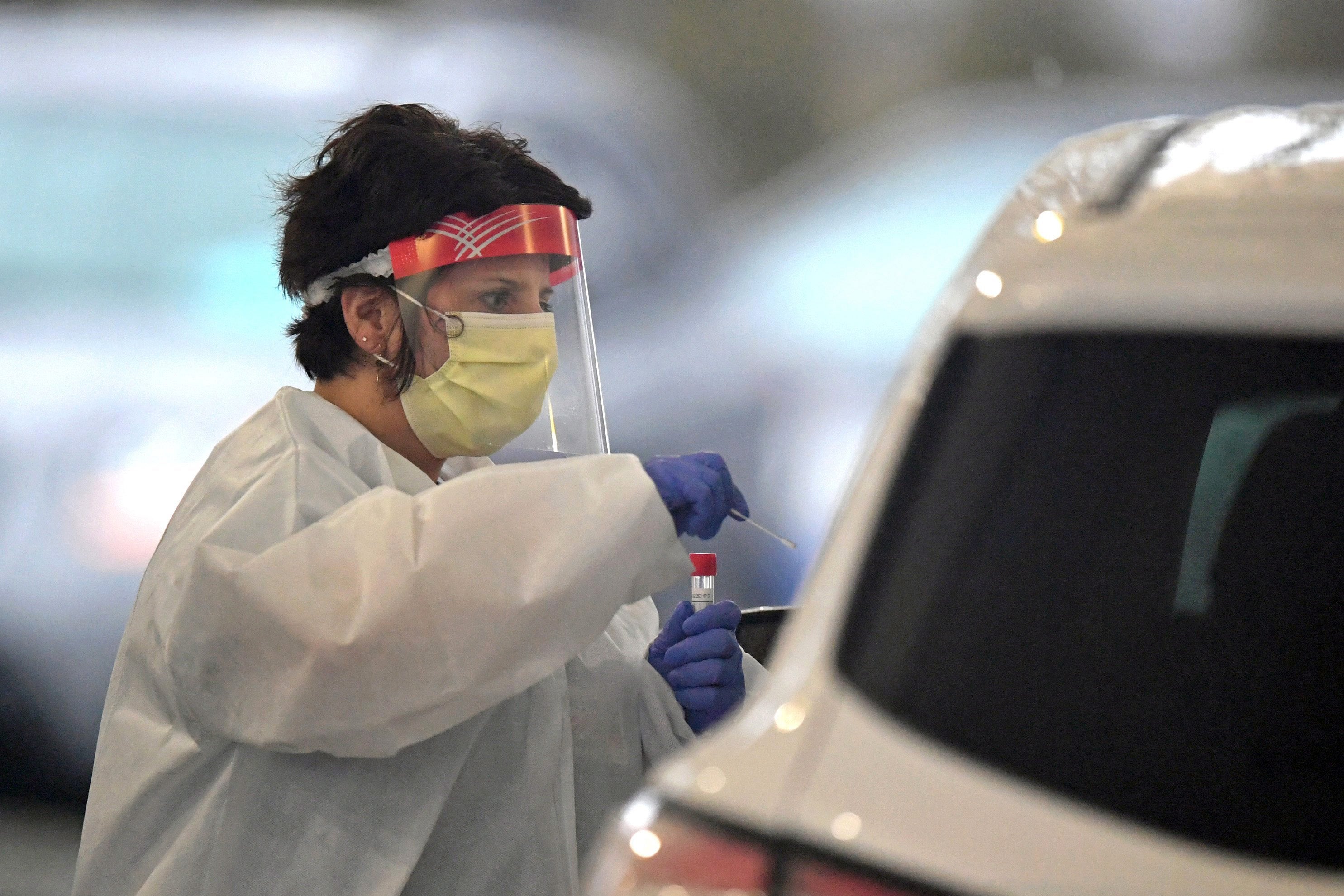 A female medical professional in a mask and other protective gear leans into a car, tube in hand.