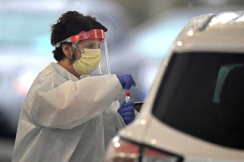 A female medical professional in a mask and other protective gear leans into a car, tube in hand.
