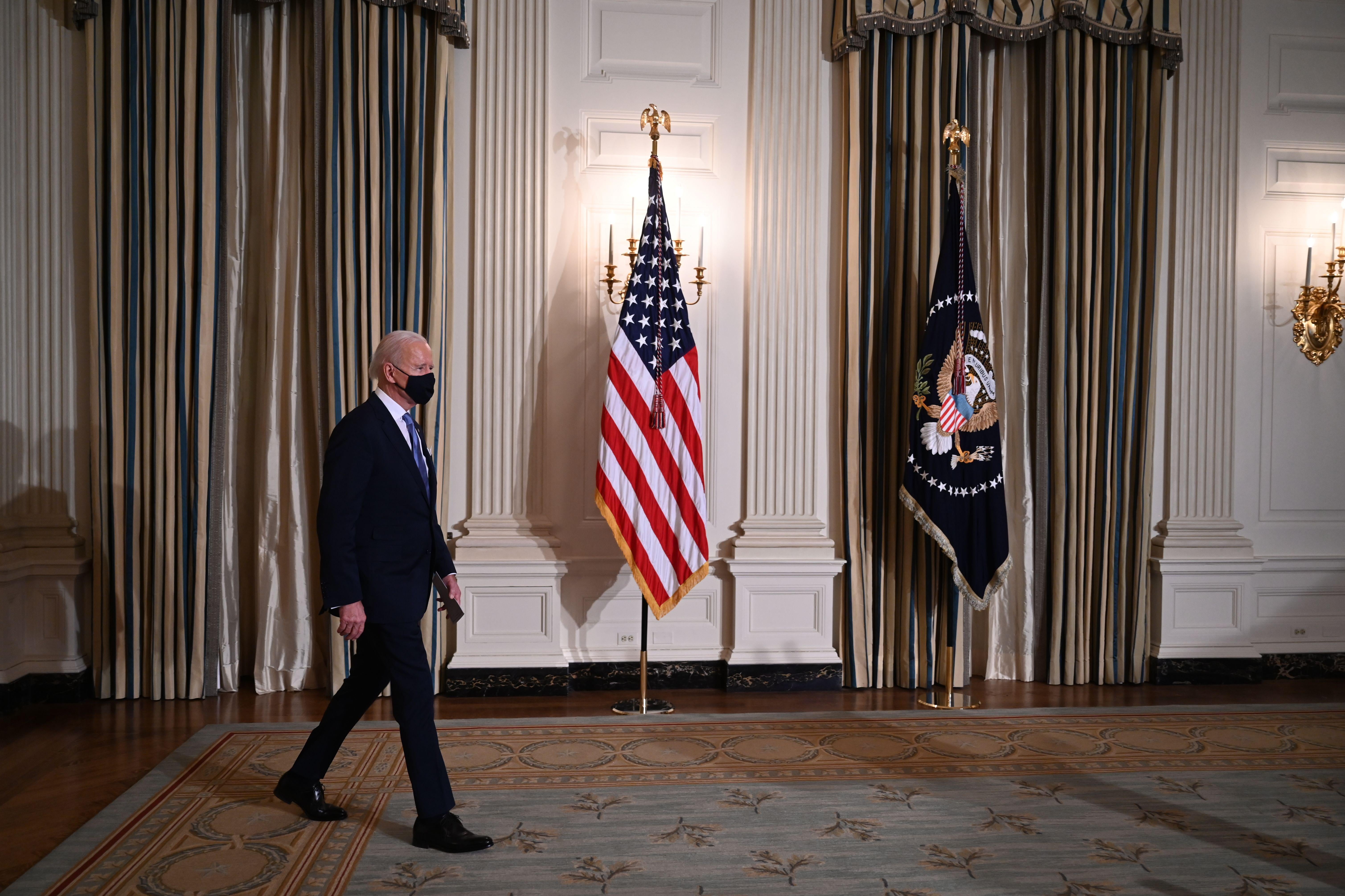 Joe Biden walks into a room in the White House with an American flag.