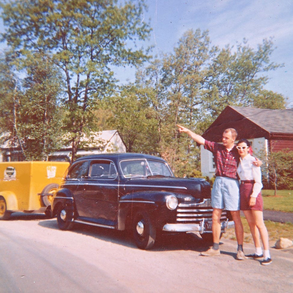 Jim and Carol stand in front of a black car. Jim points up and to the left.