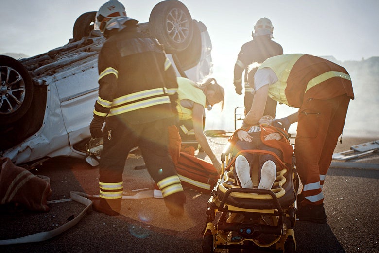 Emergency personel attend to a person in a stretcher besides a wrecked car.