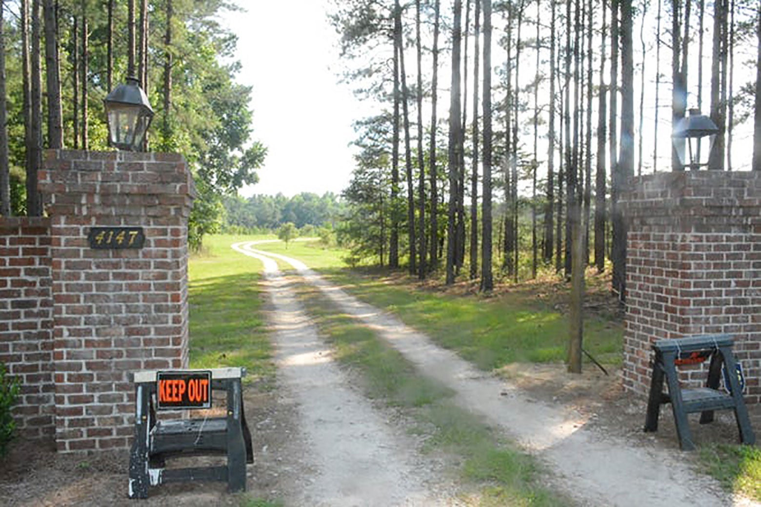 Two sawhorses bearing KEEP OUT signs are seen in front of a brick gate, through which a dirt road is seen.